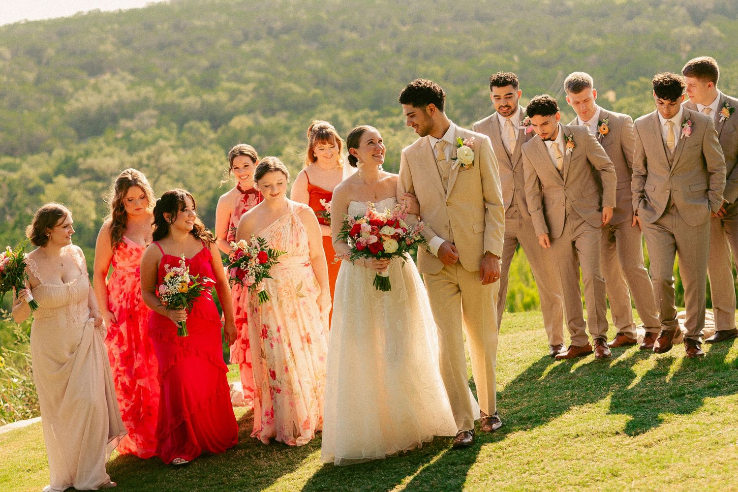 Wedding party poses outdoors with bride and groom centered, bridesmaids in red and groomsmen in tan suits, green hills behind.