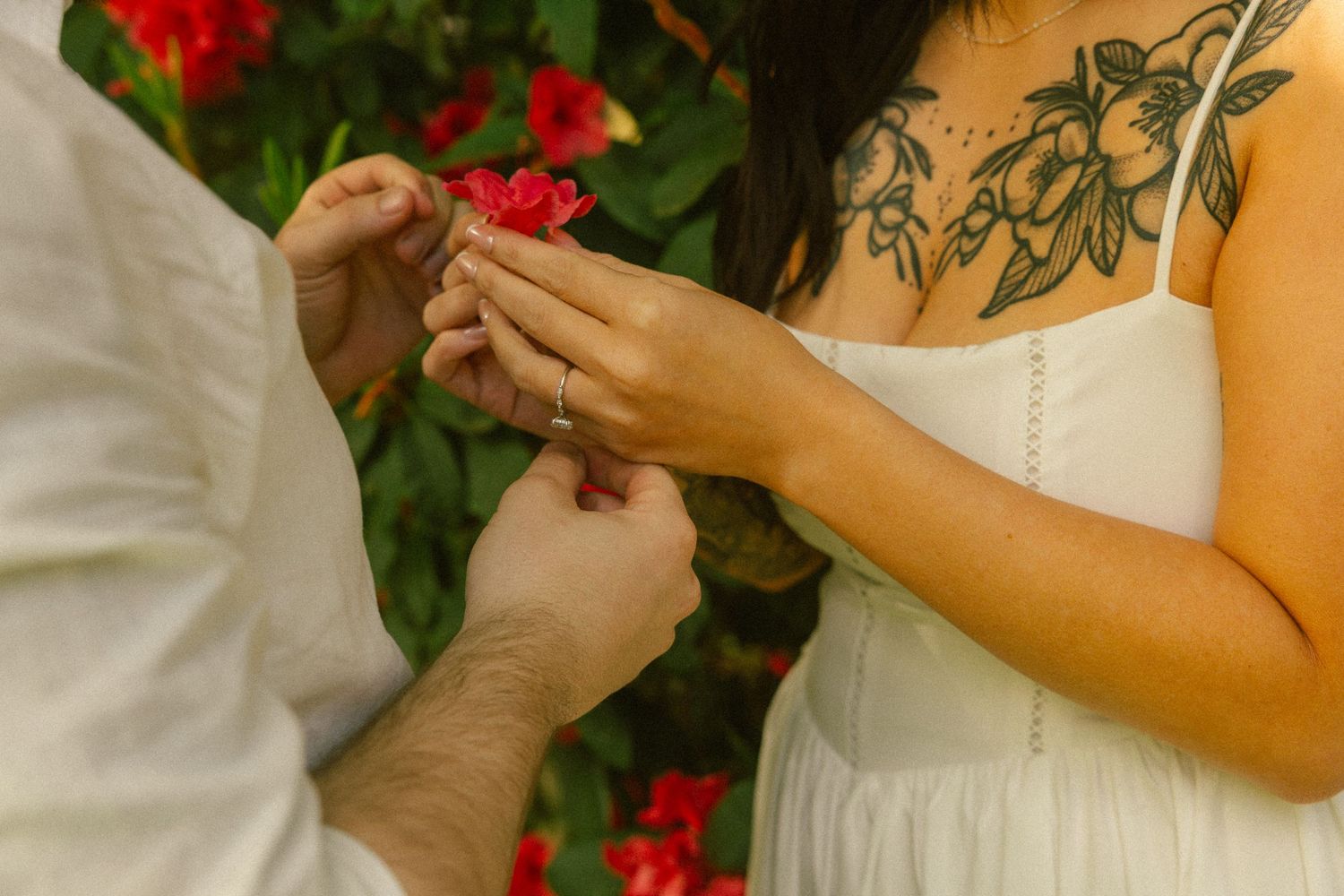 Man placing a ring on a tattooed woman's finger near red flowers during an outdoor engagement ceremony.
