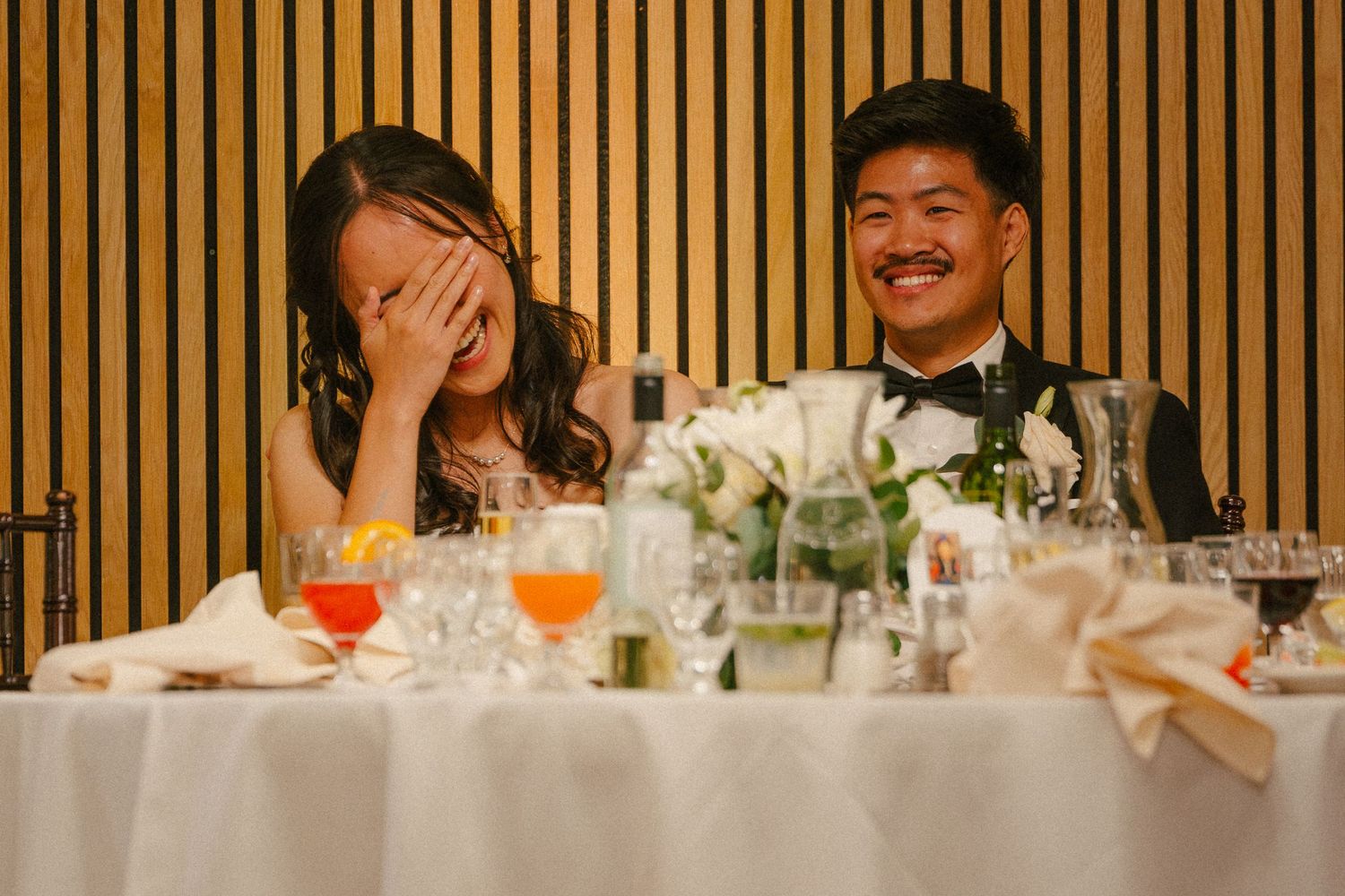 Bride laughing and covering her face while groom smiles during wedding reception speeches at elegant dinner table.
