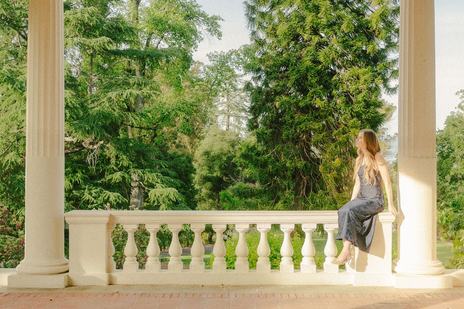 Woman in elegant dress sitting on white balustrade of classical colonnade overlooking lush green garden.