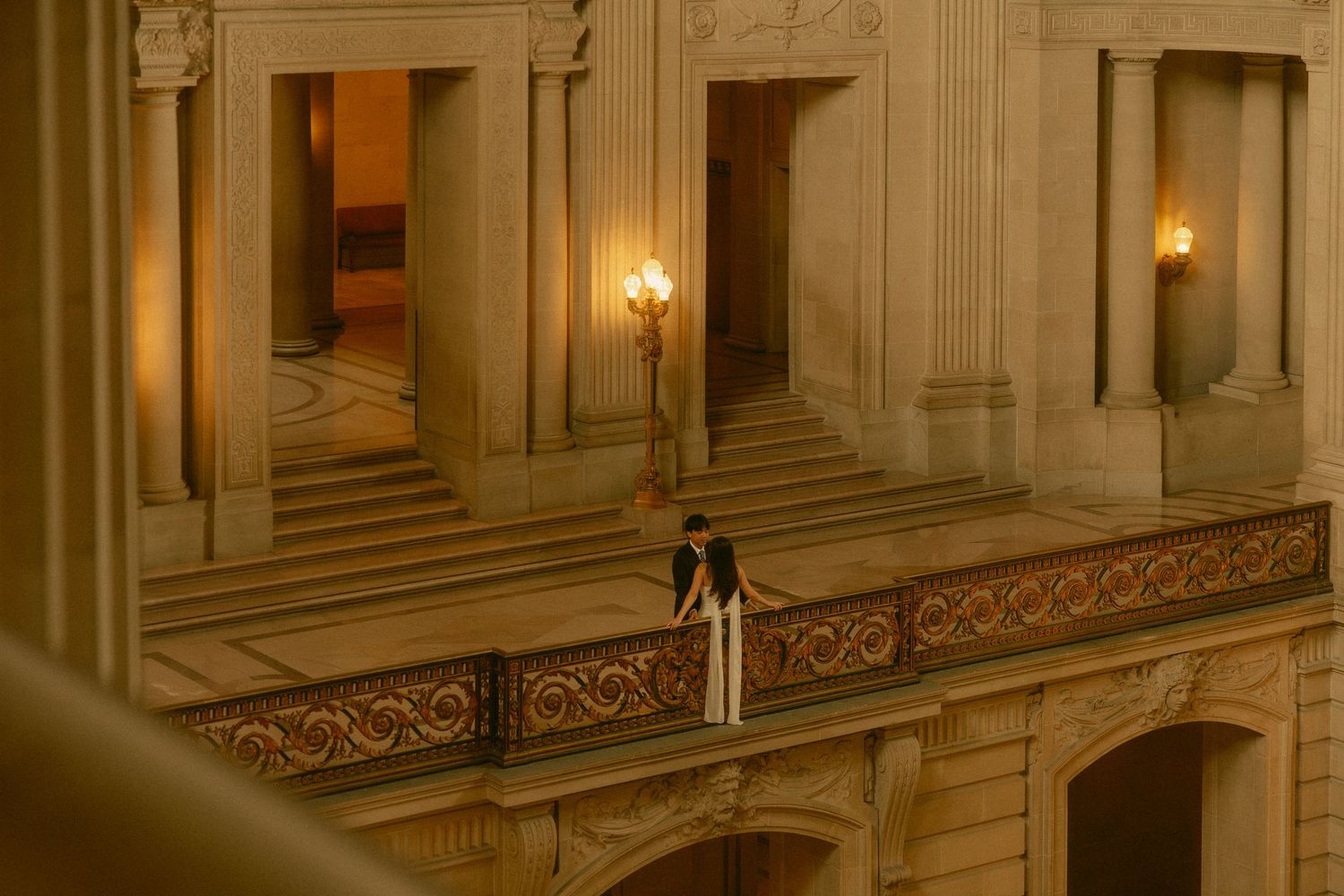 Couple sharing a romantic moment on a balcony inside an ornate golden-lit government building.