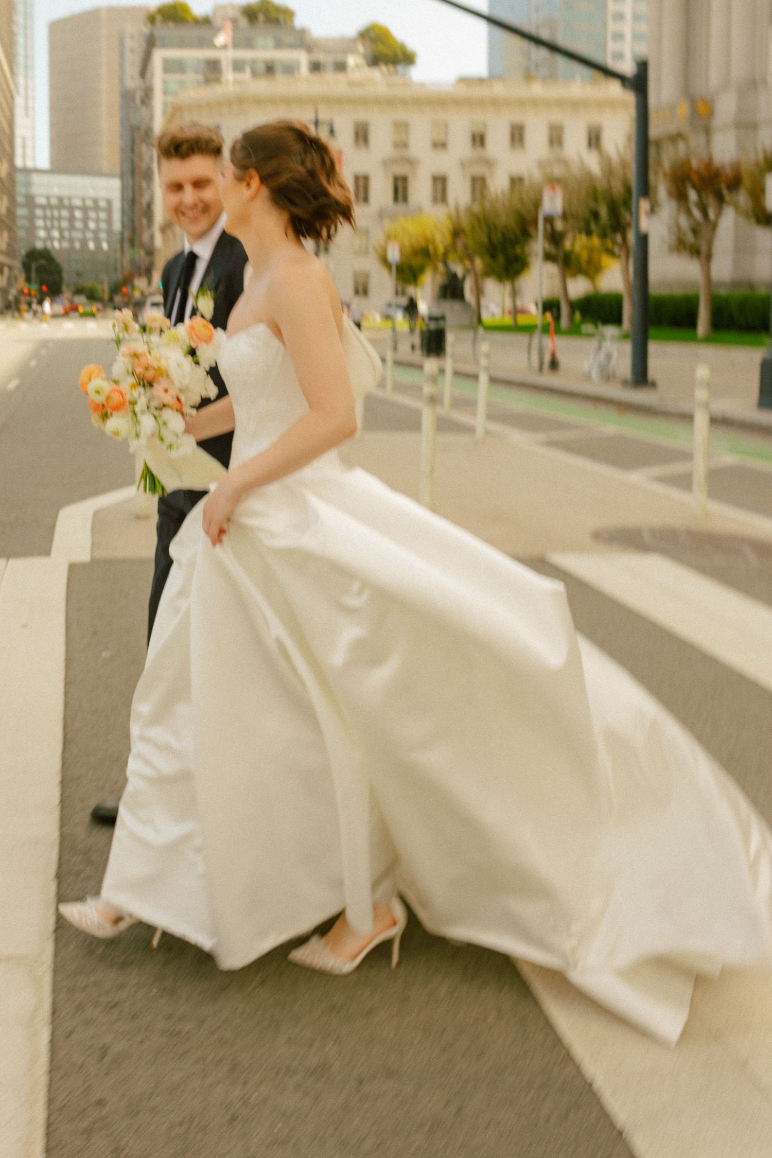 Bride in white strapless gown holding floral bouquet crosses street with groom in black suit in urban setting.