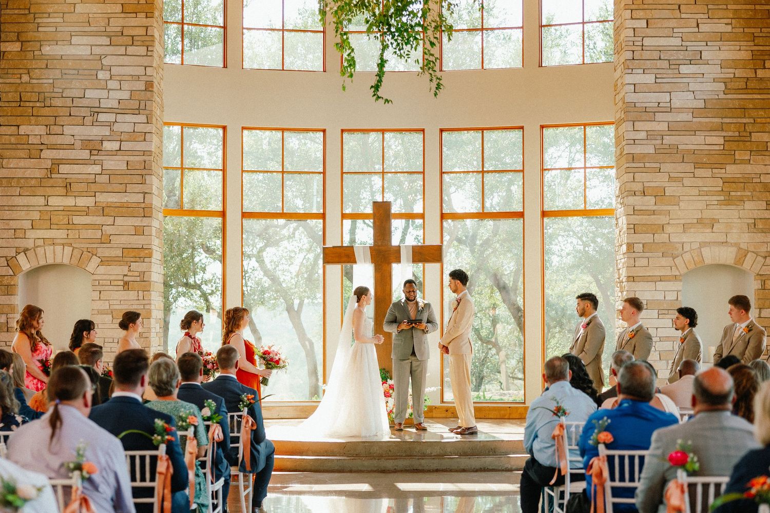 Elegant wedding ceremony in a stone chapel with large windows, wooden cross, bridal party, and seated guests.