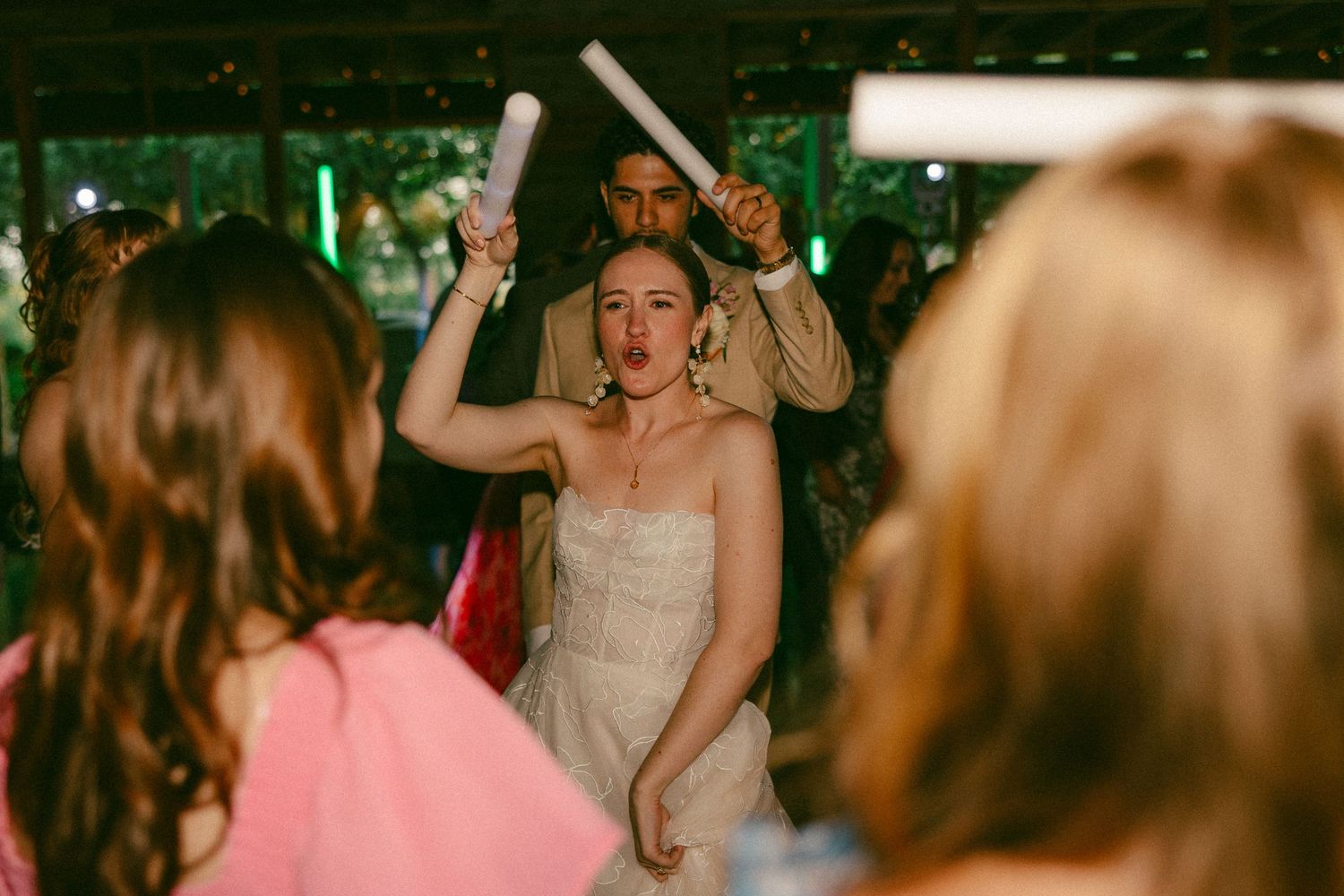 Bride in white dress dancing at wedding reception while guest holds confetti poppers overhead.