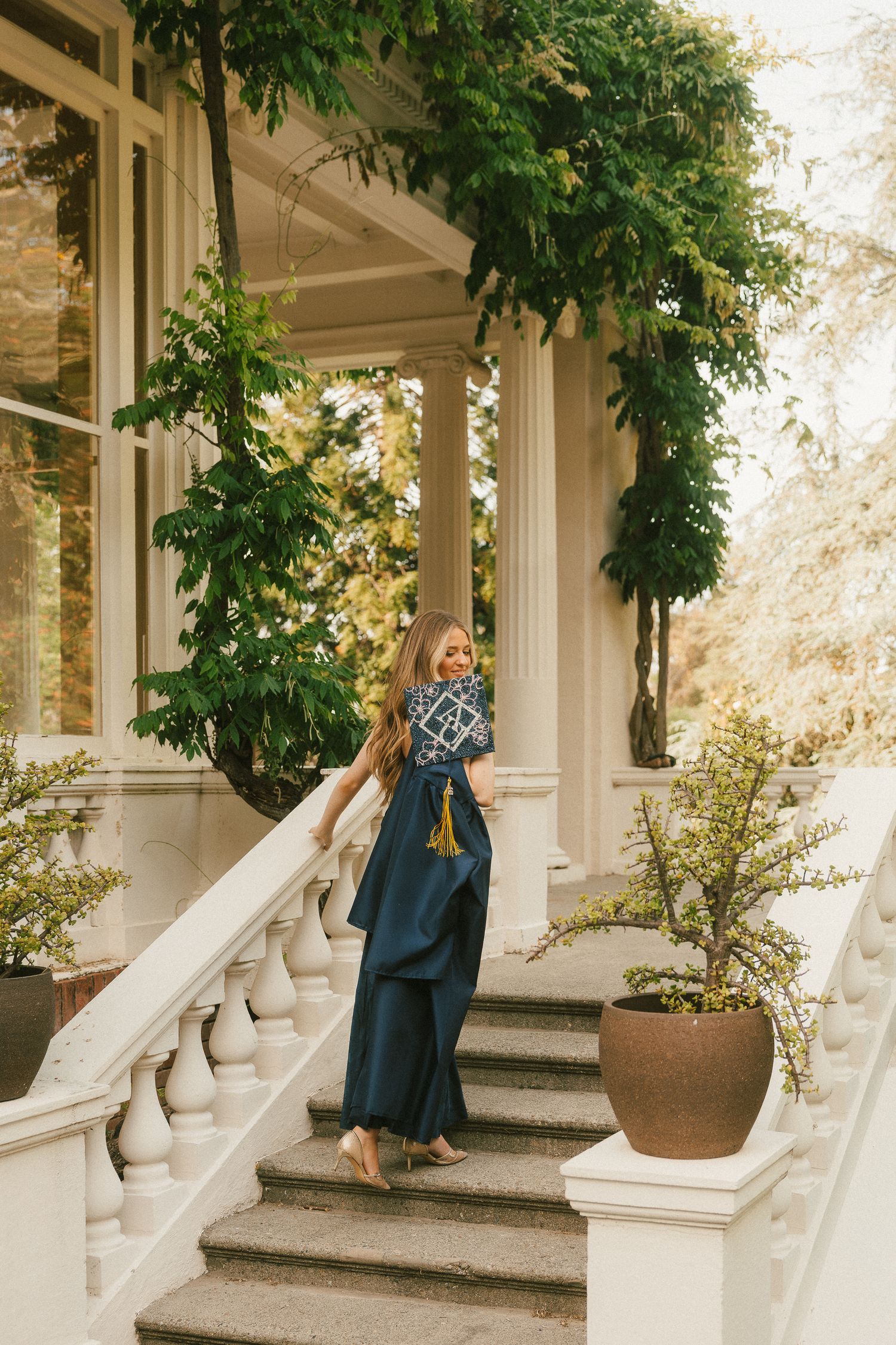 Woman in teal gown descending white balustrade stairs of elegant mansion with lush green ivy backdrop.