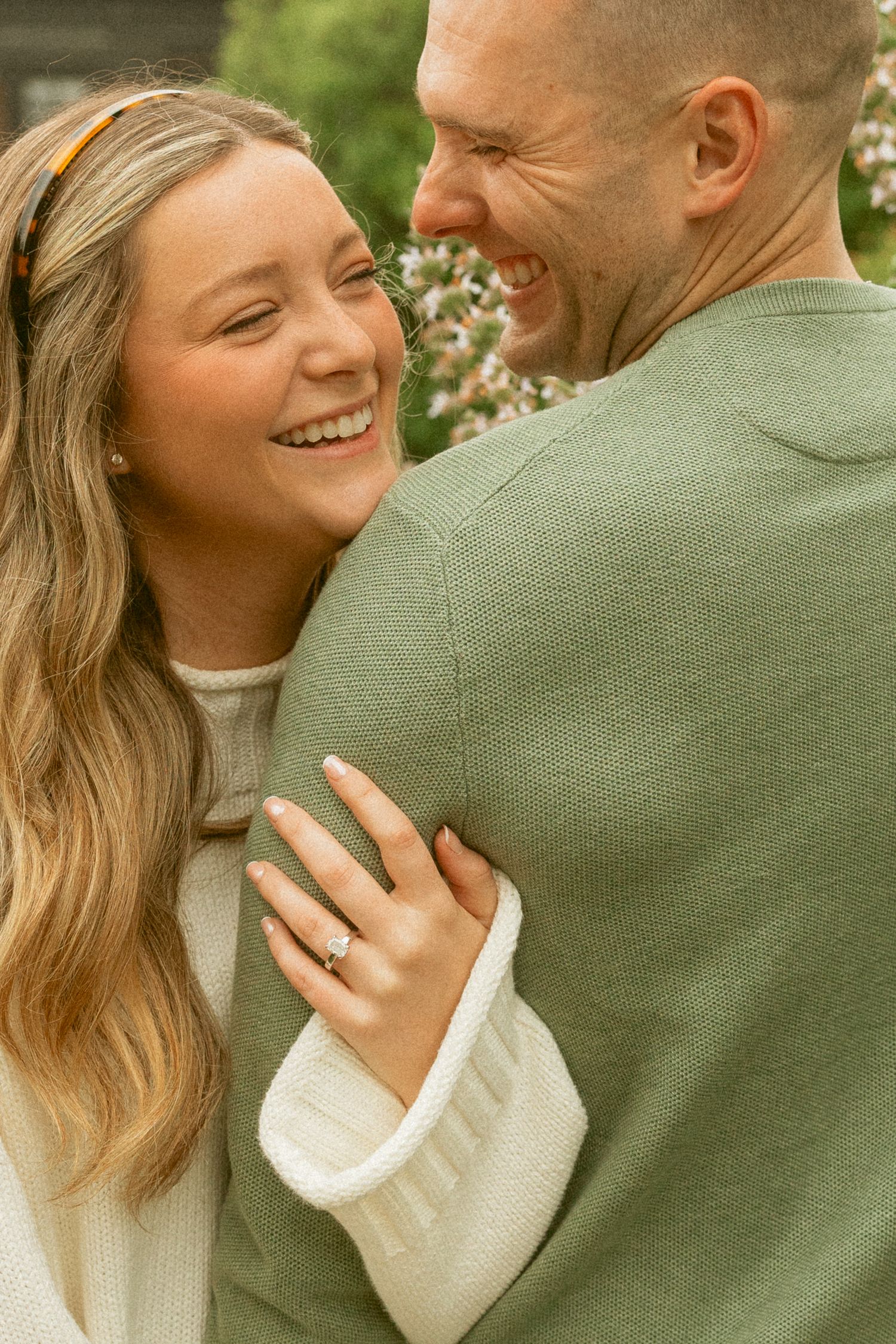 Happy couple embracing outdoors, woman wearing engagement ring, smiling joyfully in green and white sweaters.