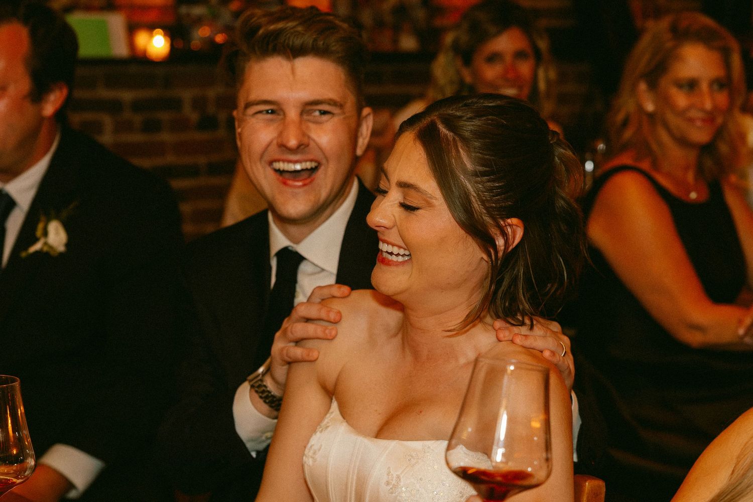 Happy couple laughing together at wedding reception, bride holding red wine glass, groom in black suit with tie.