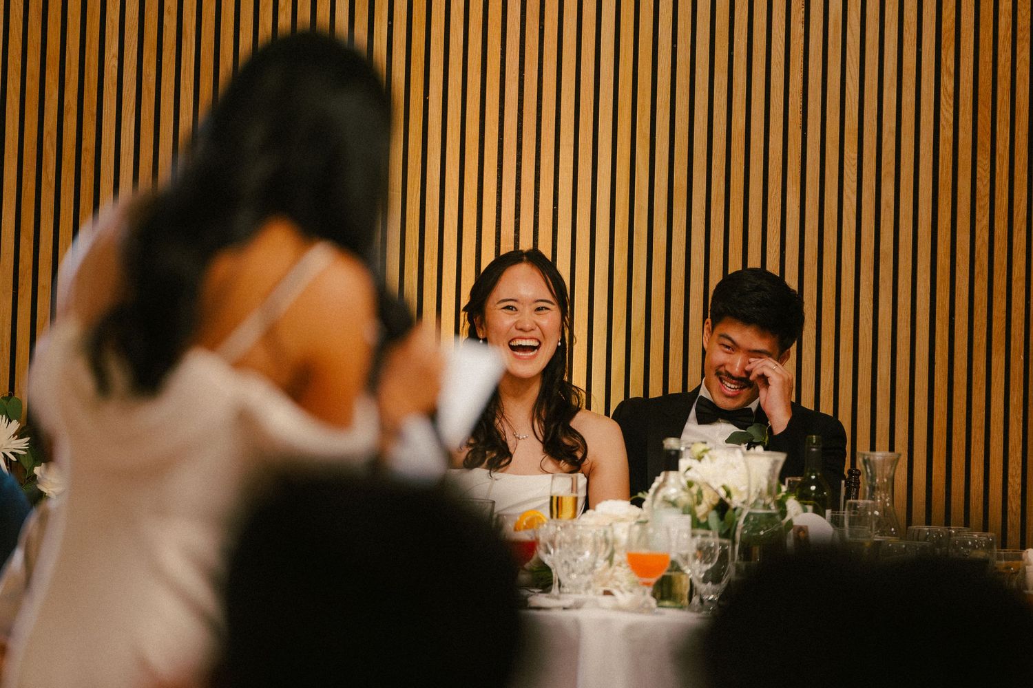 Bride and groom laughing joyfully at wedding reception dinner table with floral centerpieces and wood panel backdrop.