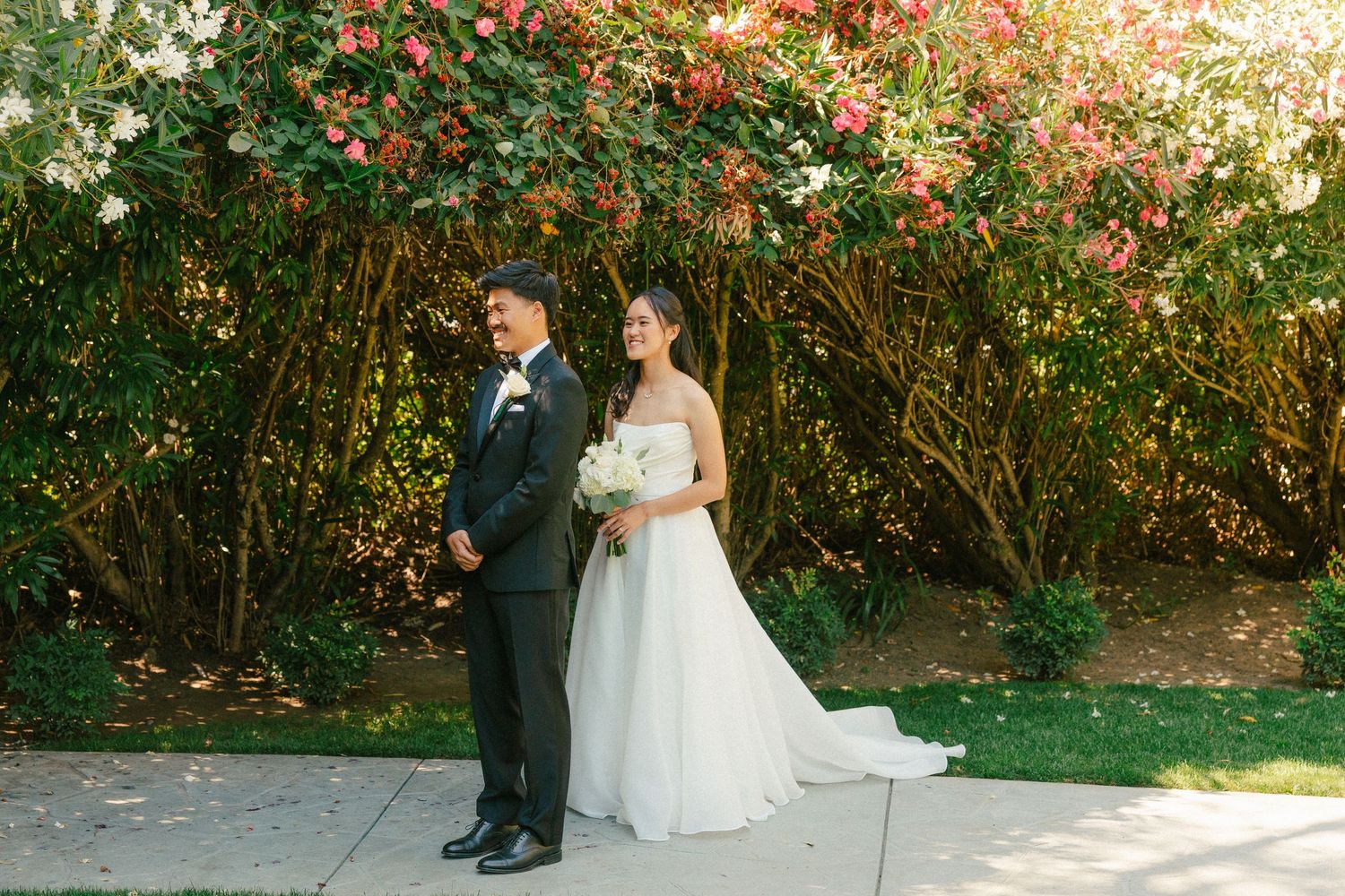 Bride and groom pose together in front of lush flowering bushes, bride holding white bouquet in elegant strapless gown.