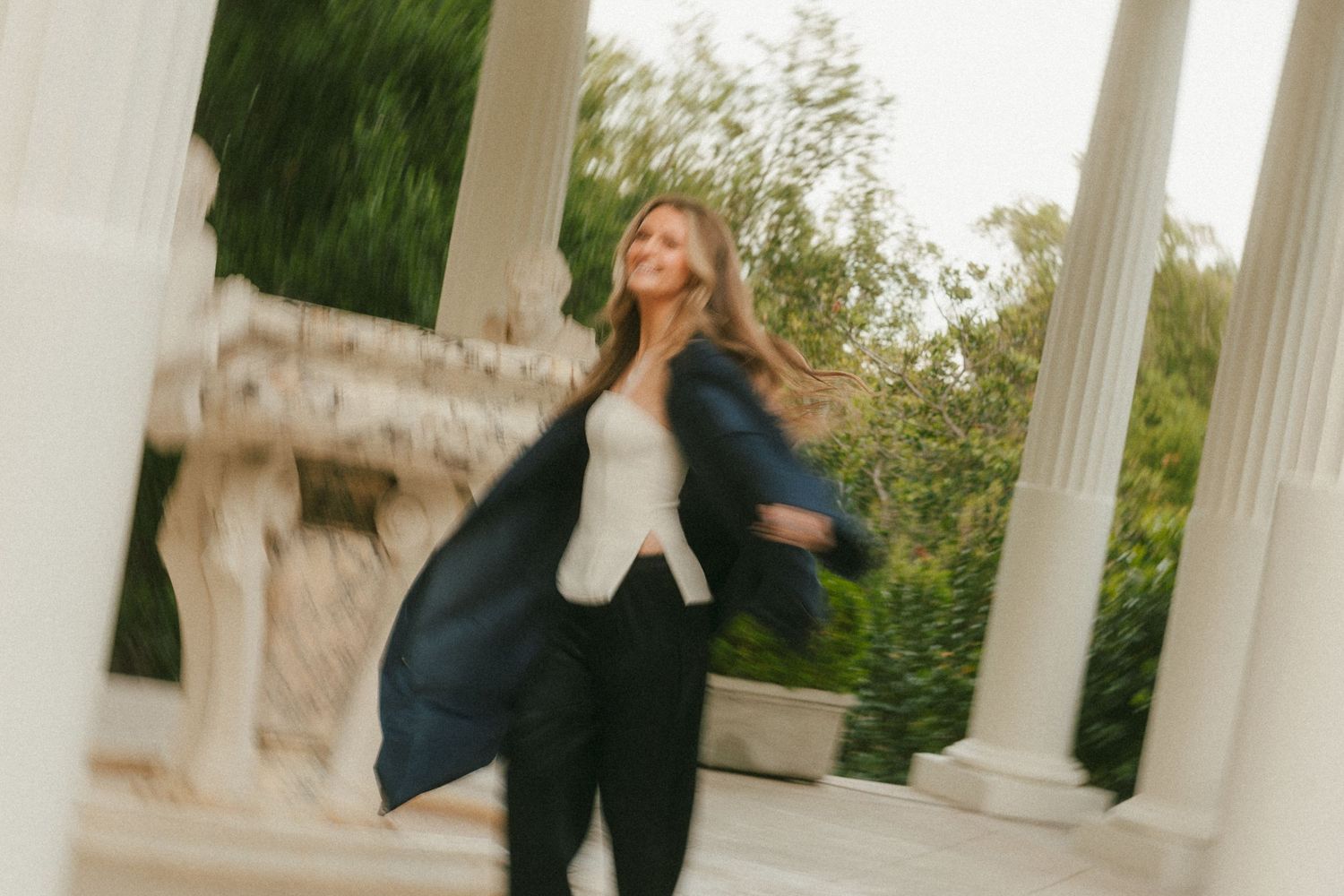Woman in dark blazer spinning joyfully among white columns and lush greenery in an elegant outdoor setting.
