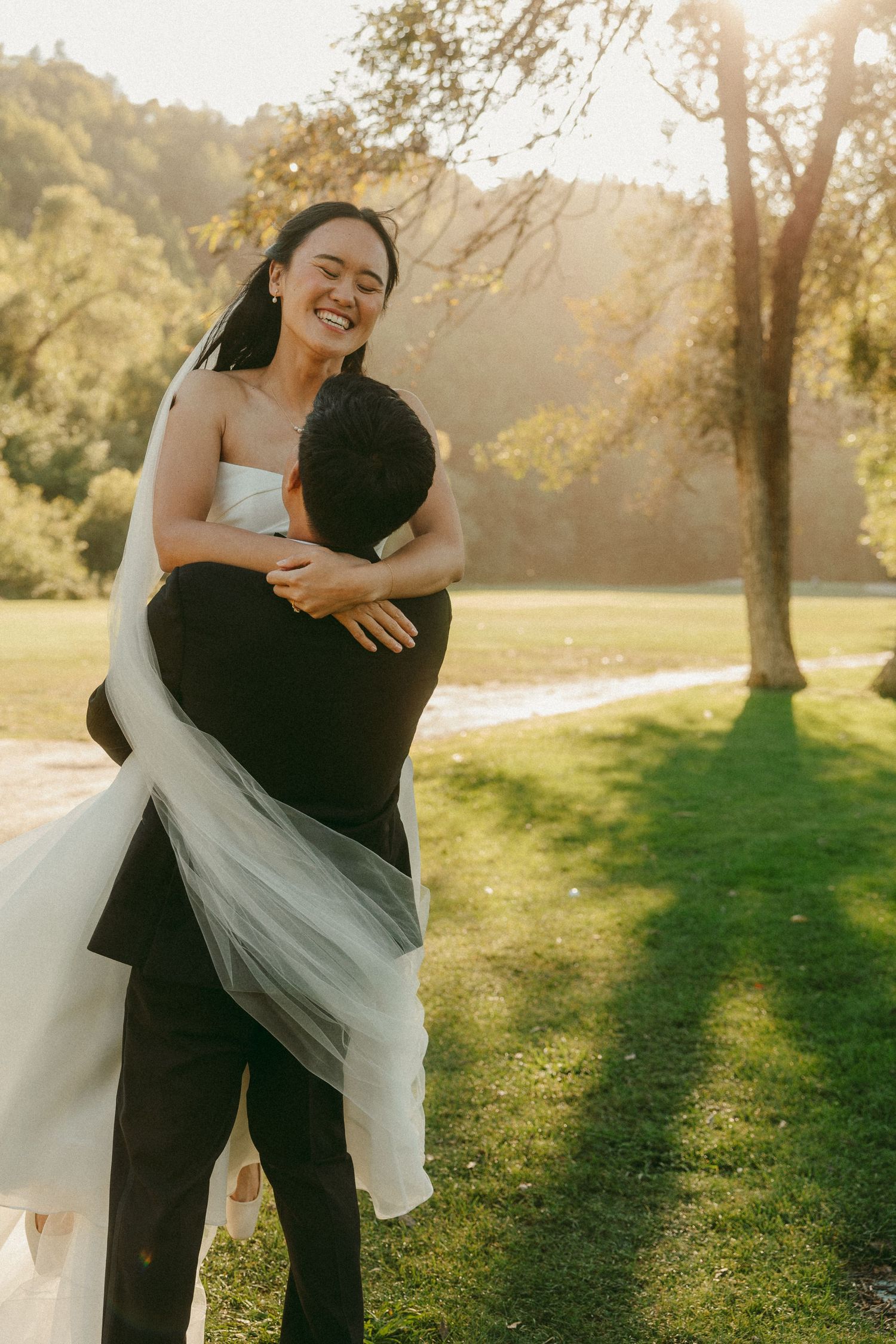 Groom lifting bride in wedding dress outdoors at golden hour on lush green lawn with sunlit trees.