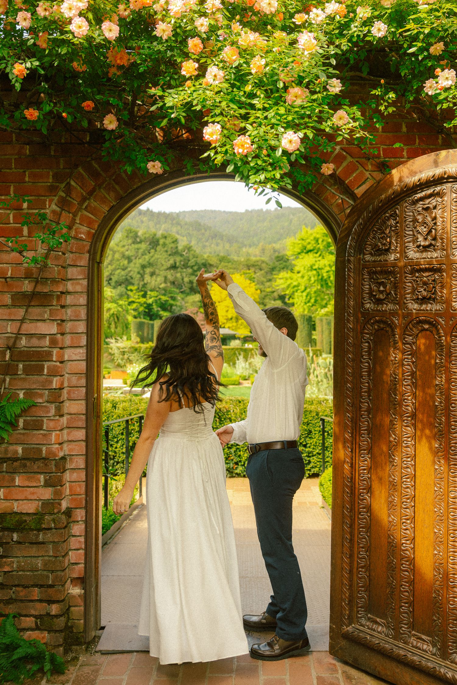 Couple dancing in elegant brick archway adorned with roses, overlooking lush green garden landscape.