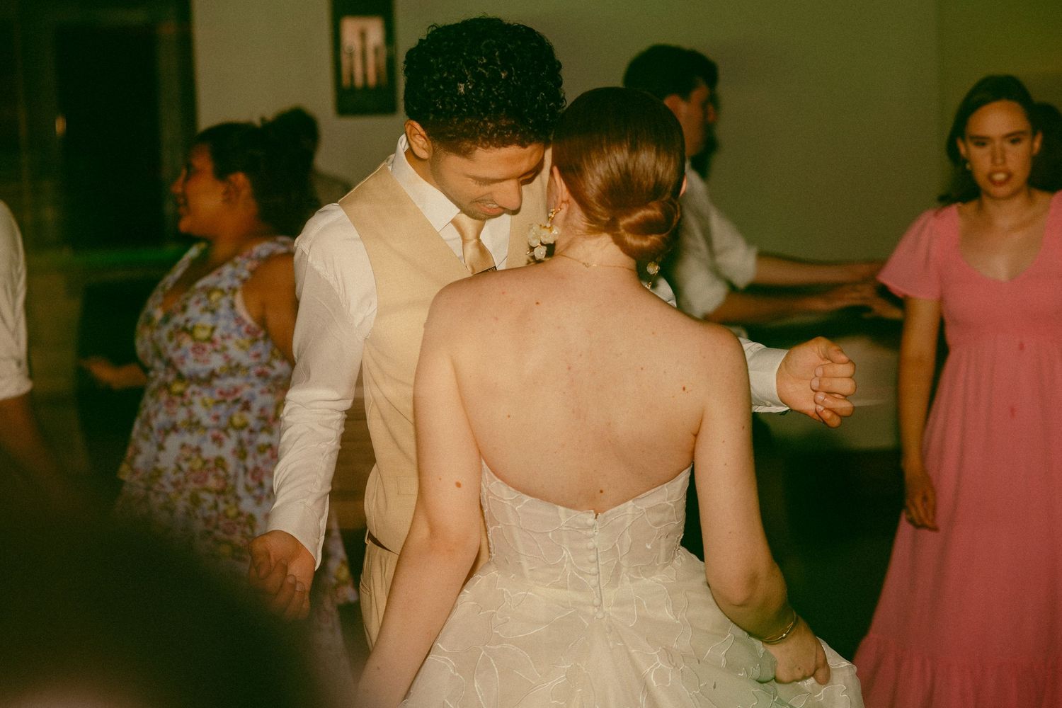 Bride and groom share first dance at wedding reception, bride in strapless gown, guests dancing in background.