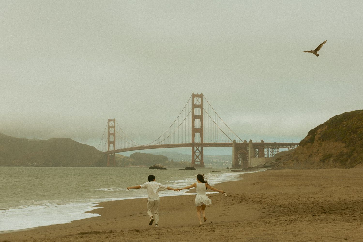 Couple holding hands running on Baker Beach with the Golden Gate Bridge in the fog behind them.
