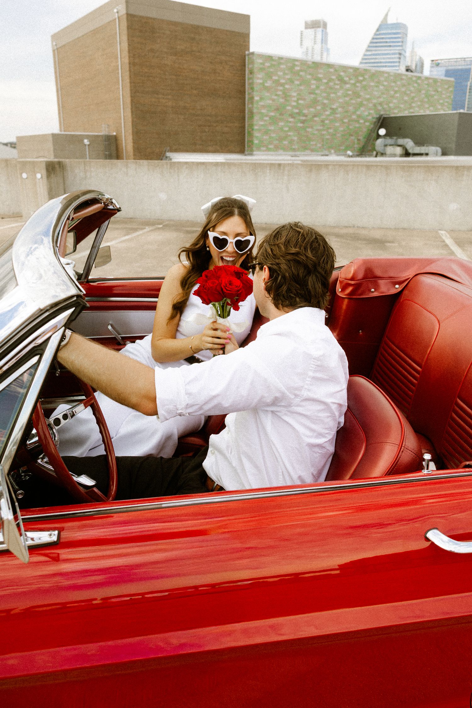 Couple sharing romantic moment in vintage red convertible car against urban backdrop.