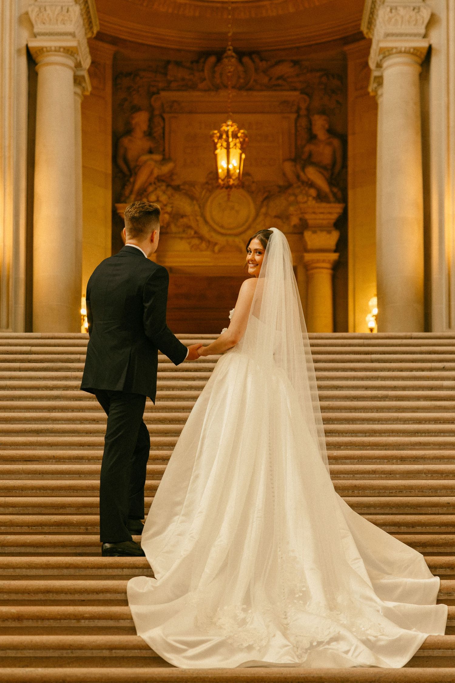 Bride and groom hold hands ascending grand marble staircase in ornate wedding venue, bride's long veil trailing behind.