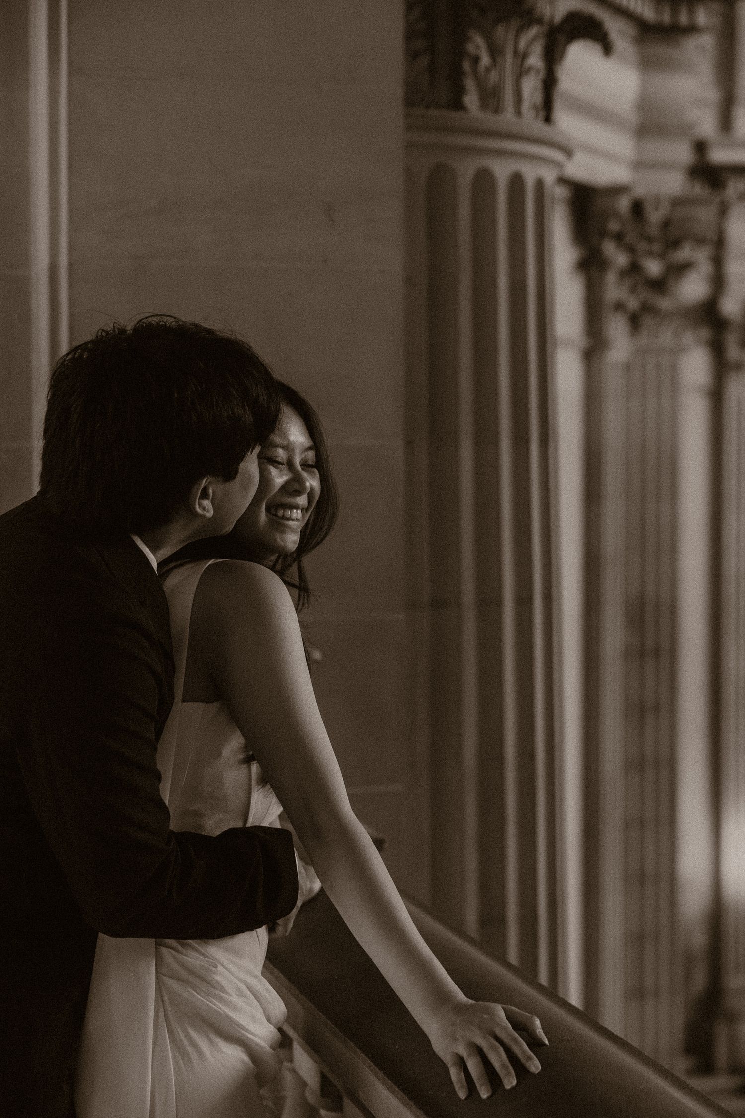 Black and white wedding photo of a smiling bride in white gown embraced by groom near ornate columns.
