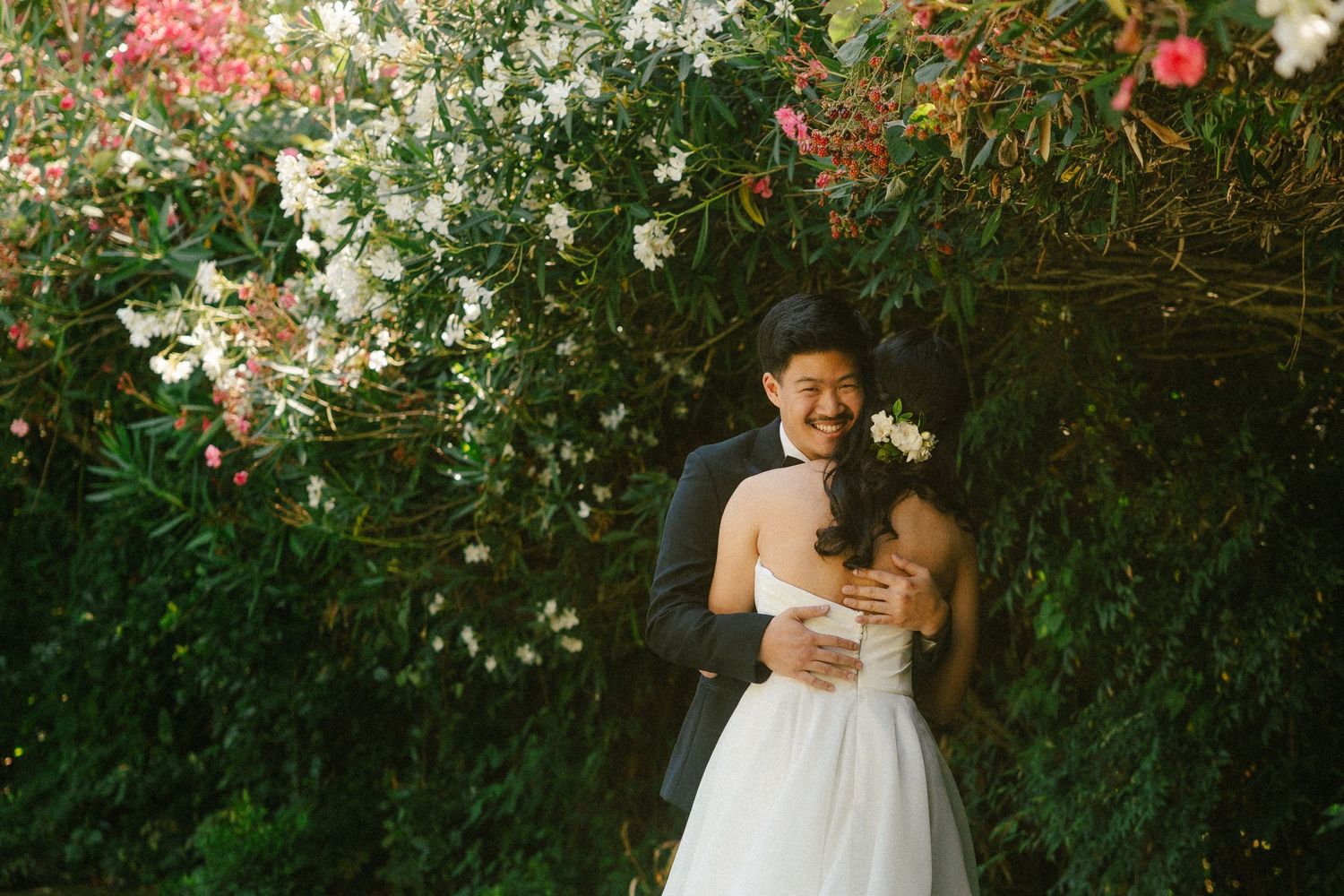 A smiling groom in a black suit embraces his bride in a white dress among lush green foliage and blooming flowers.