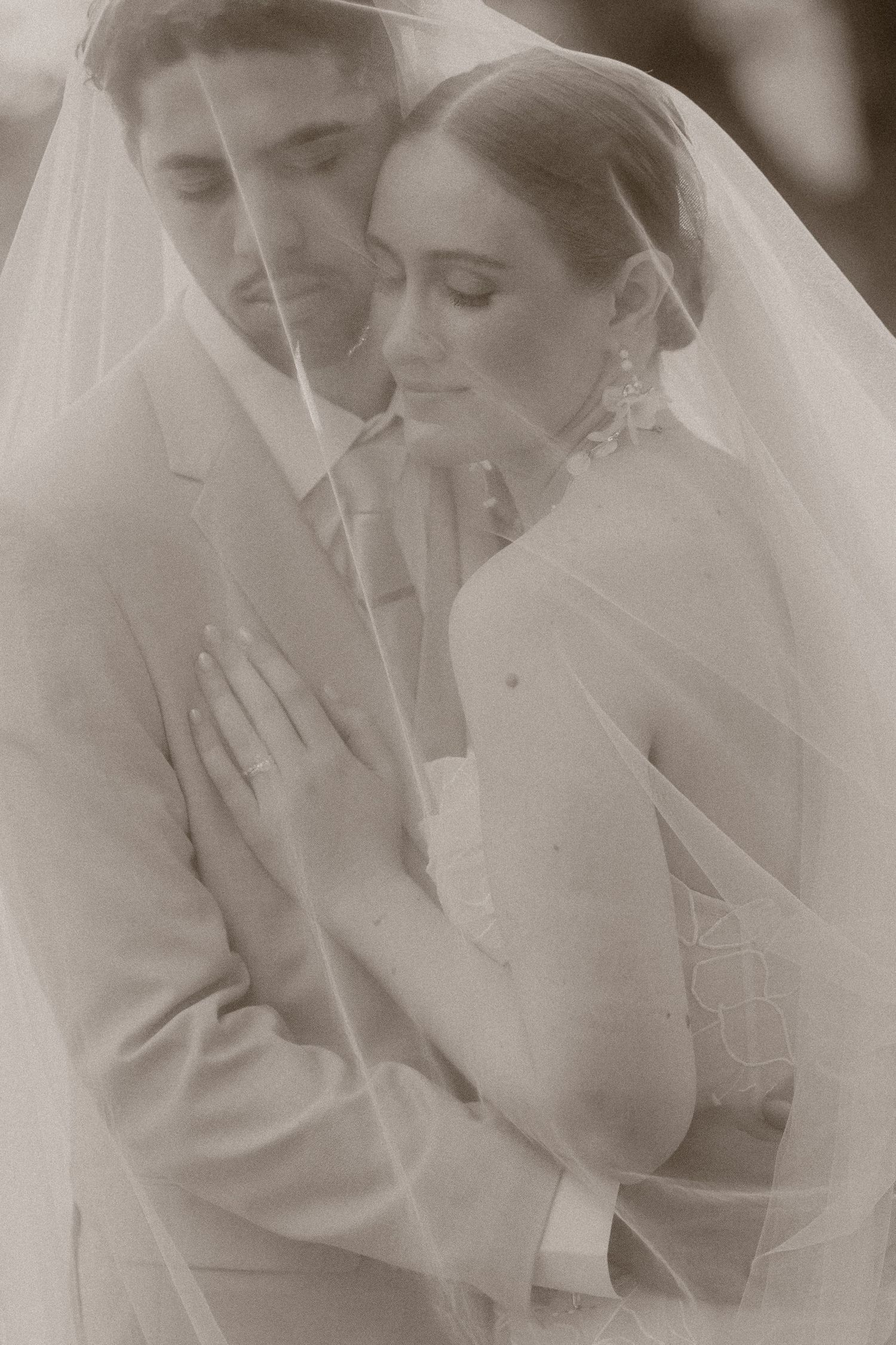 Bride and groom embracing tenderly under a wedding veil in a romantic sepia-toned portrait.