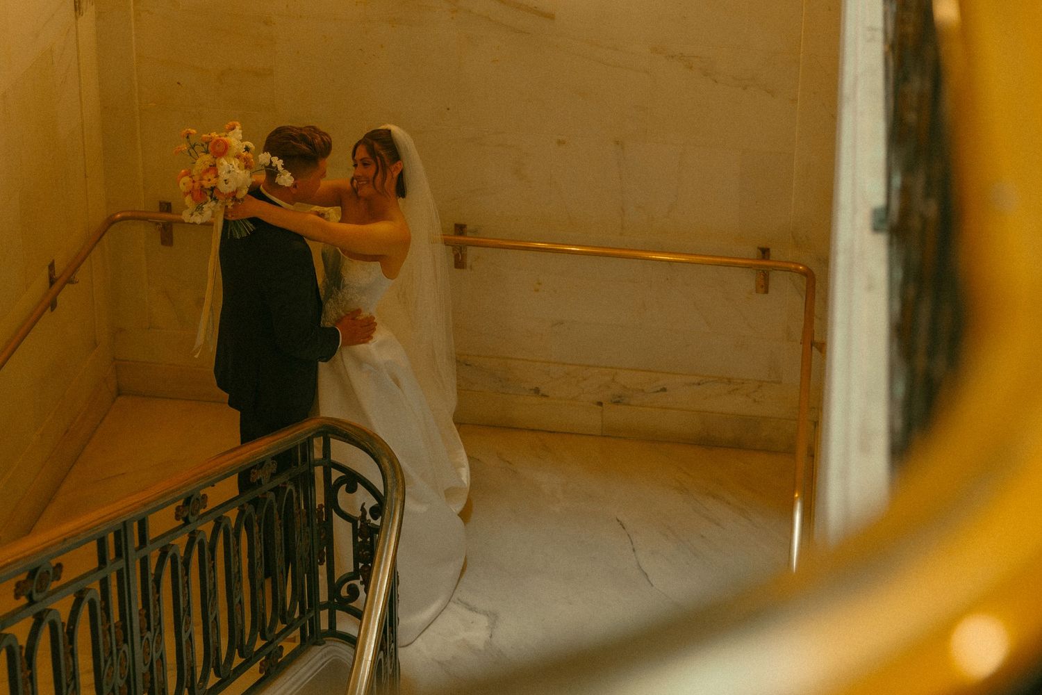 Bride and groom share romantic embrace on elegant marble staircase with gold railing during wedding ceremony.