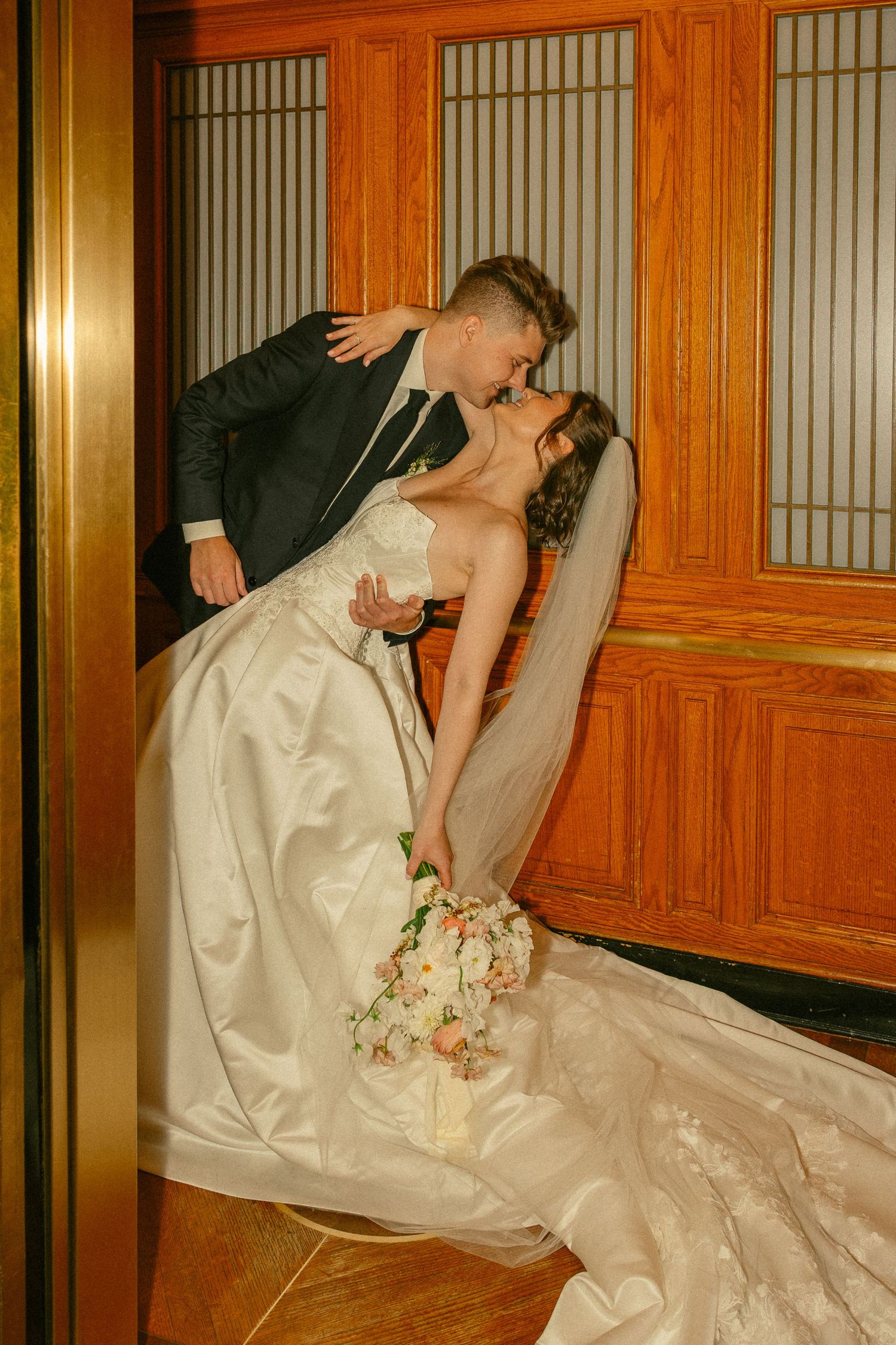 A bride and groom share a romantic dip kiss in a elegant wood-paneled venue, her white gown and bouquet trailing gracefully.