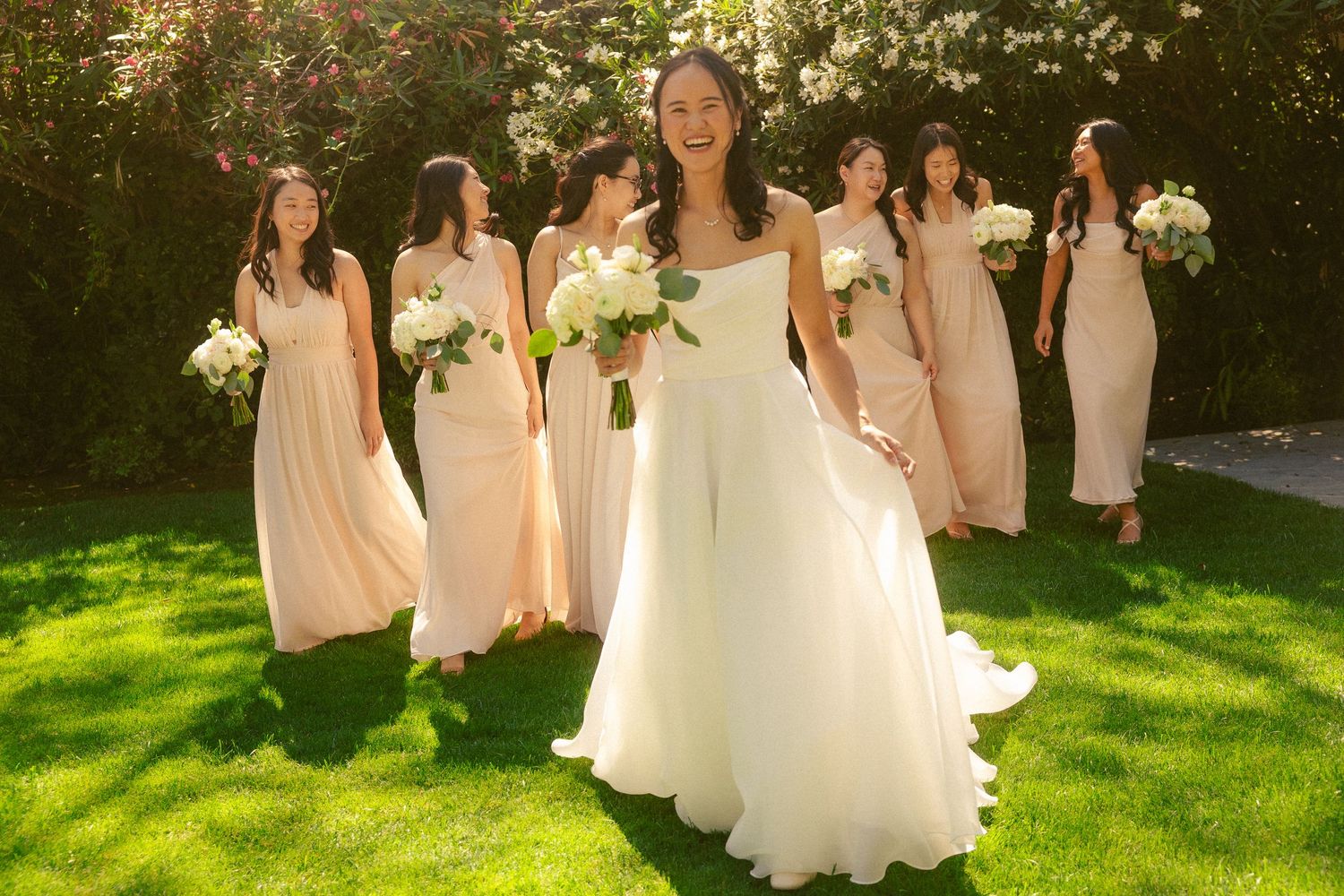 Bride in white gown laughs with bridesmaids in blush pink dresses holding white bouquets on green lawn.