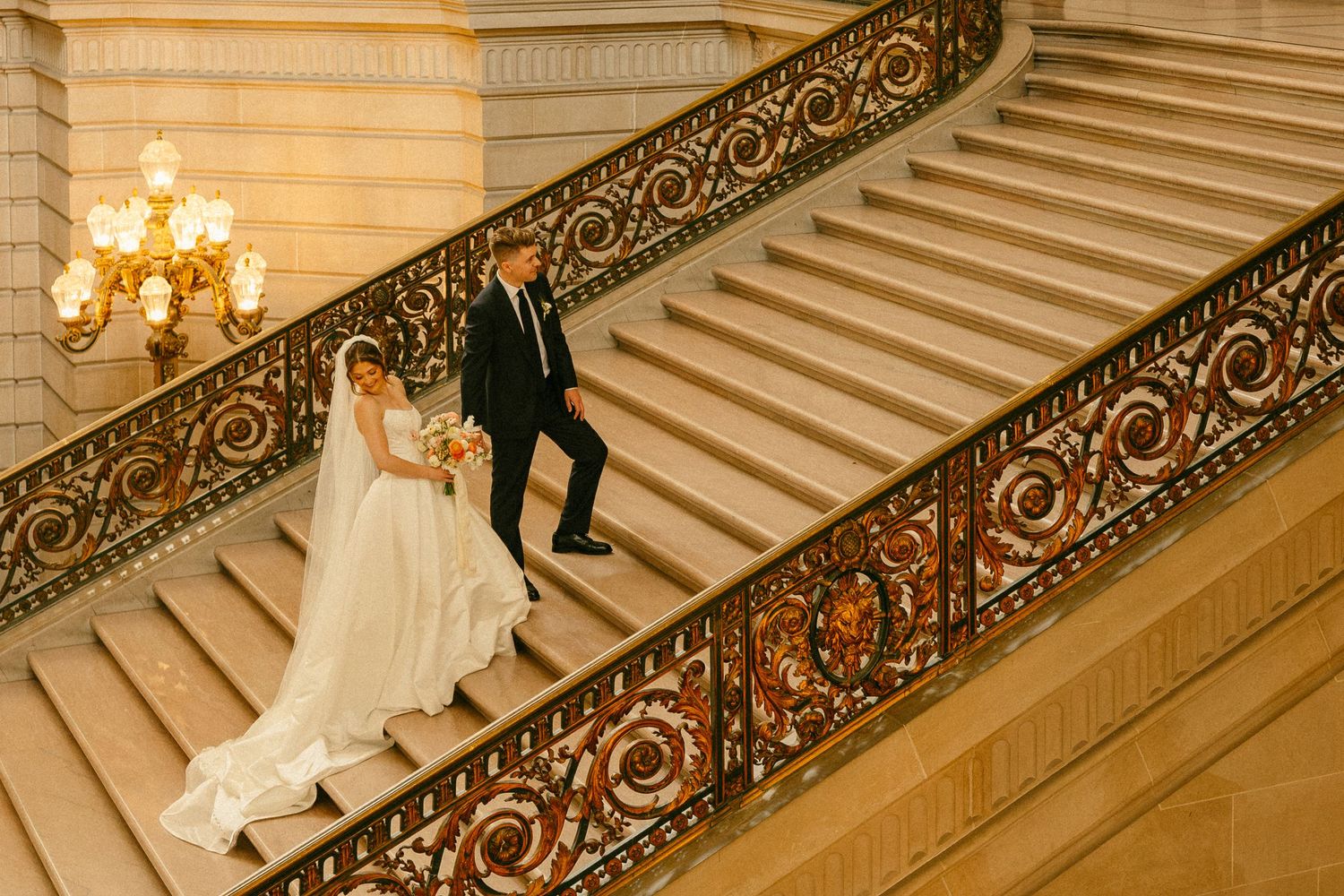 Bride and groom descend ornate grand staircase with gilded railings in elegant wedding venue.