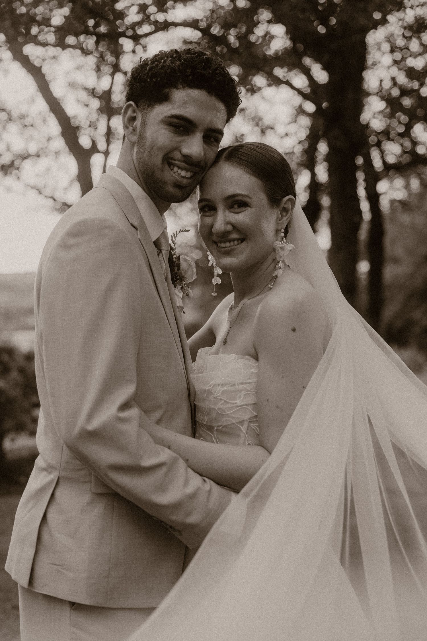 Sepia-toned wedding portrait of smiling bride in veil and groom in tan suit embracing outdoors among trees.