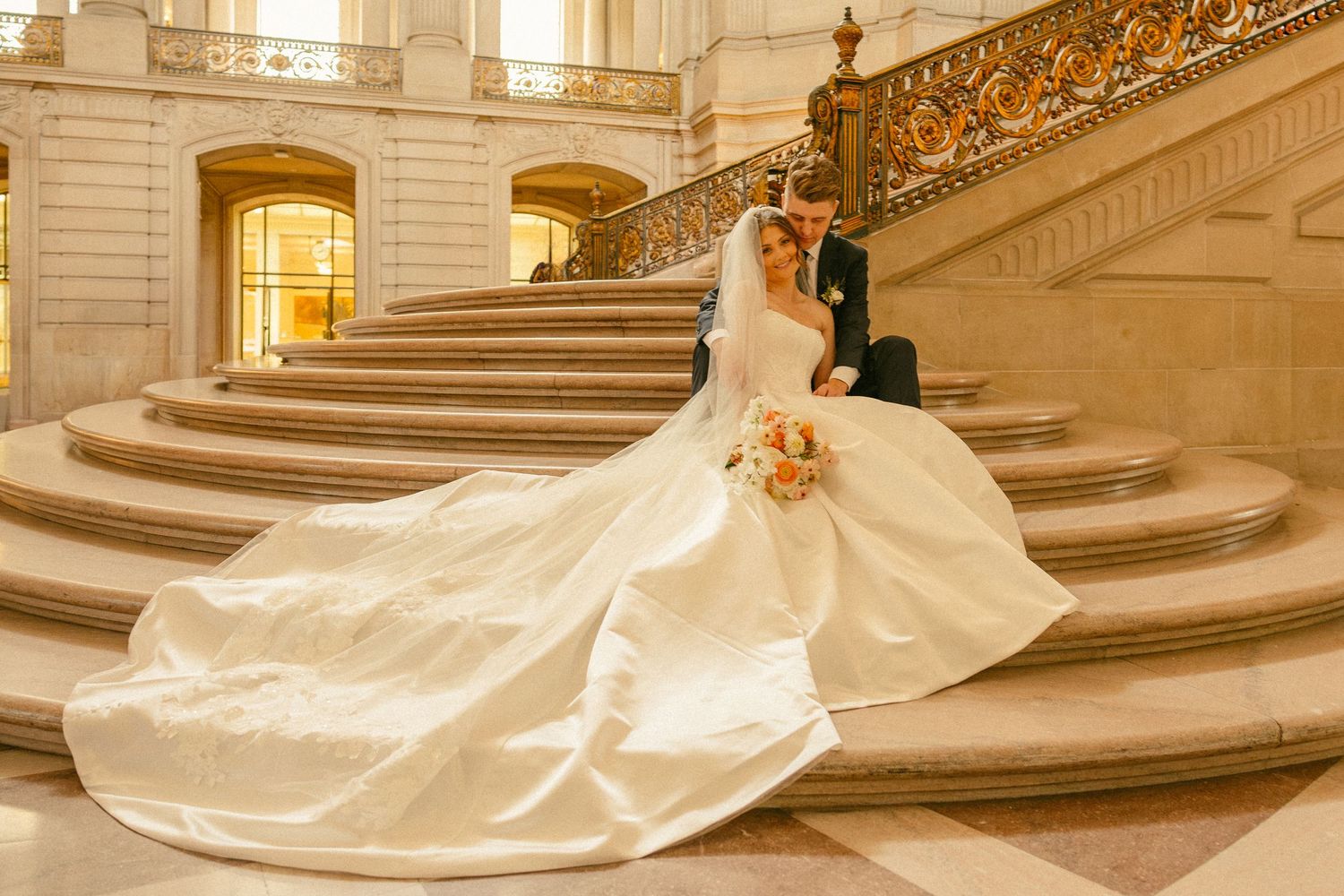 Bride and groom sitting on grand marble staircase, bride in elegant white gown with long train, holding bouquet.