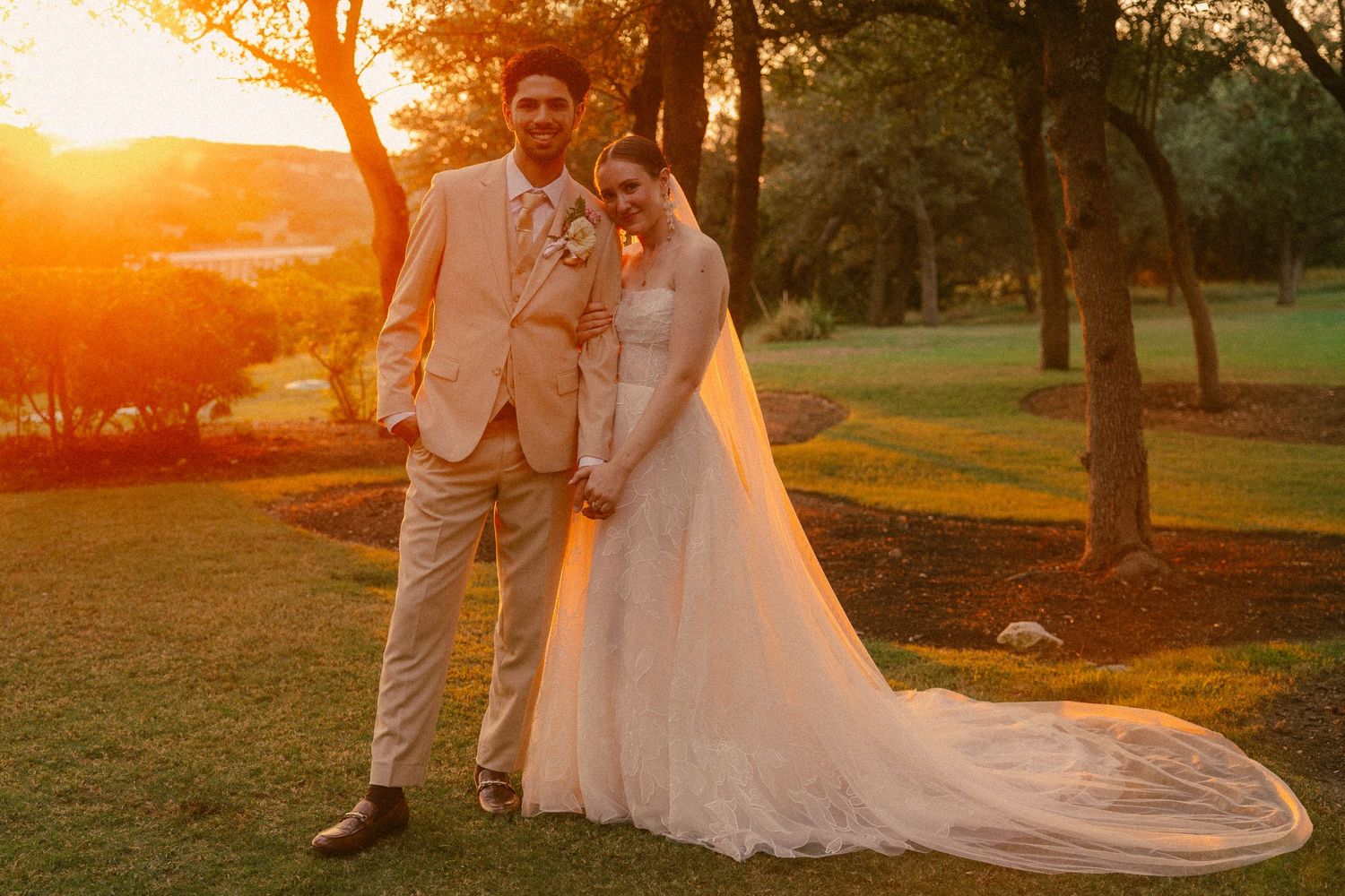 Bride and groom holding hands at golden sunset on golf course, bride in long white gown with veil, groom in tan suit.