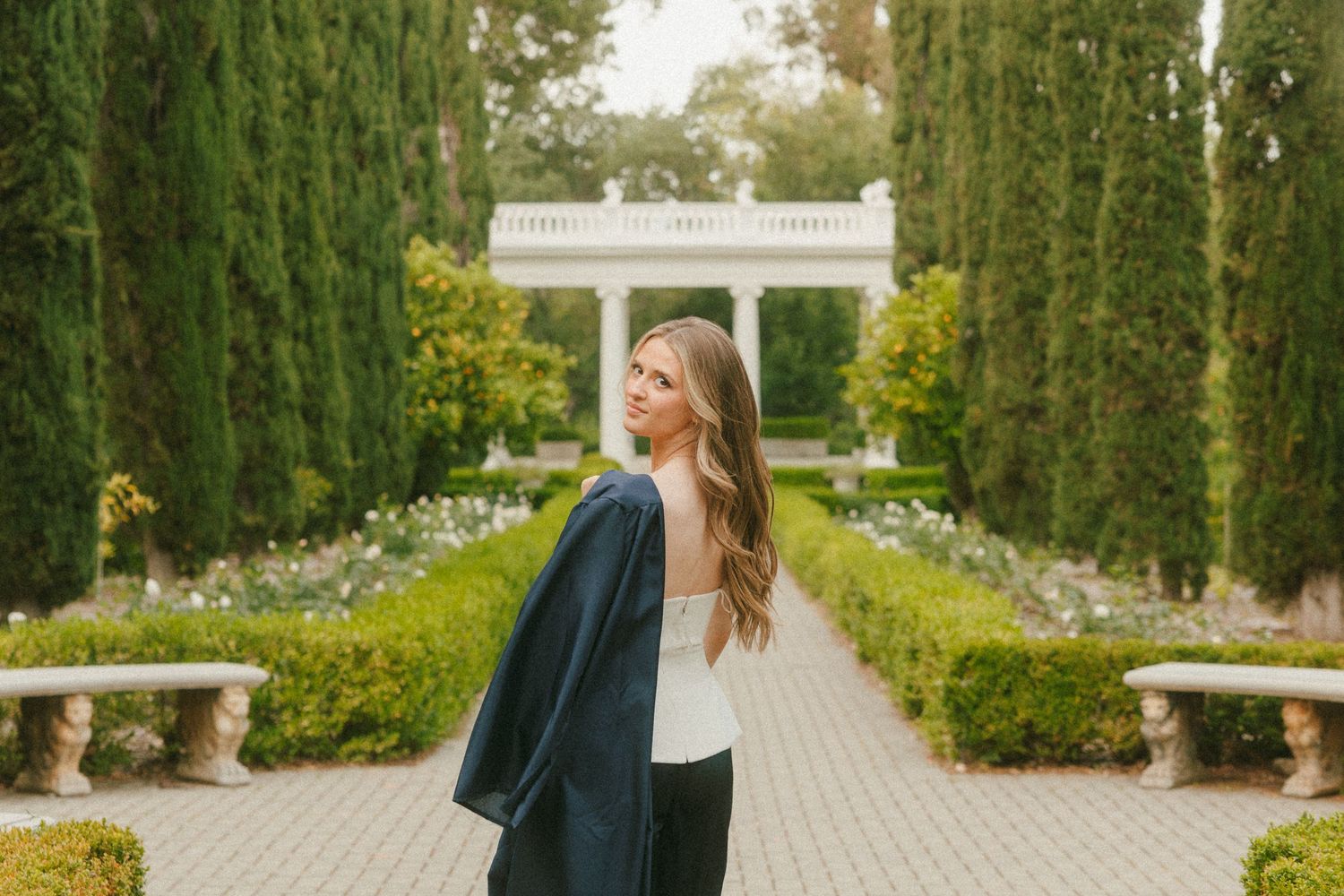 Woman in graduation gown poses in elegant formal garden with tall hedges and white pergola backdrop.