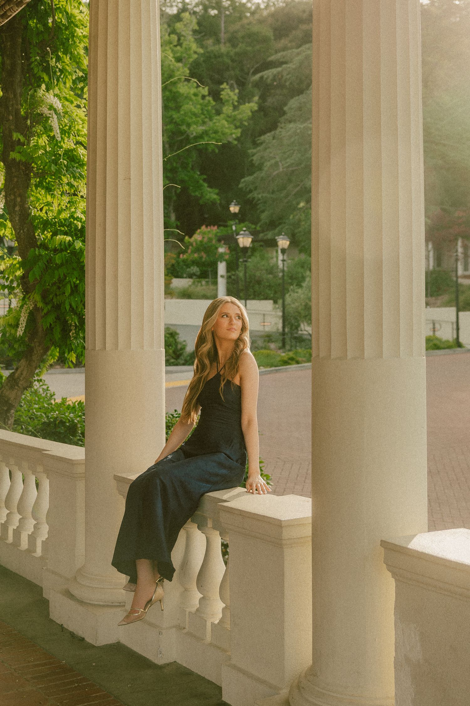 Young woman in black dress sitting on white balustrade between classical columns in a sunlit outdoor setting.