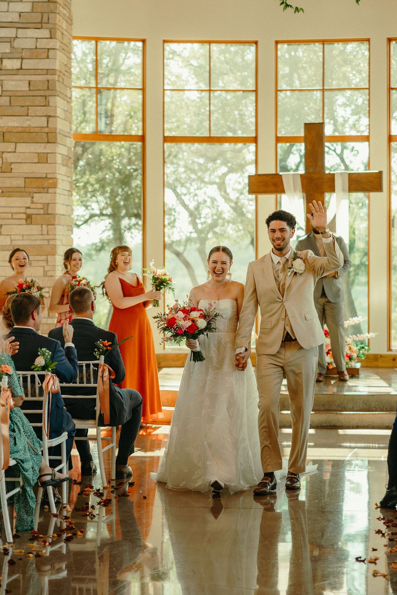 Joyful newlyweds walk down the aisle in a sunlit chapel, bride holding a bouquet, groom waving, guests cheering around them.