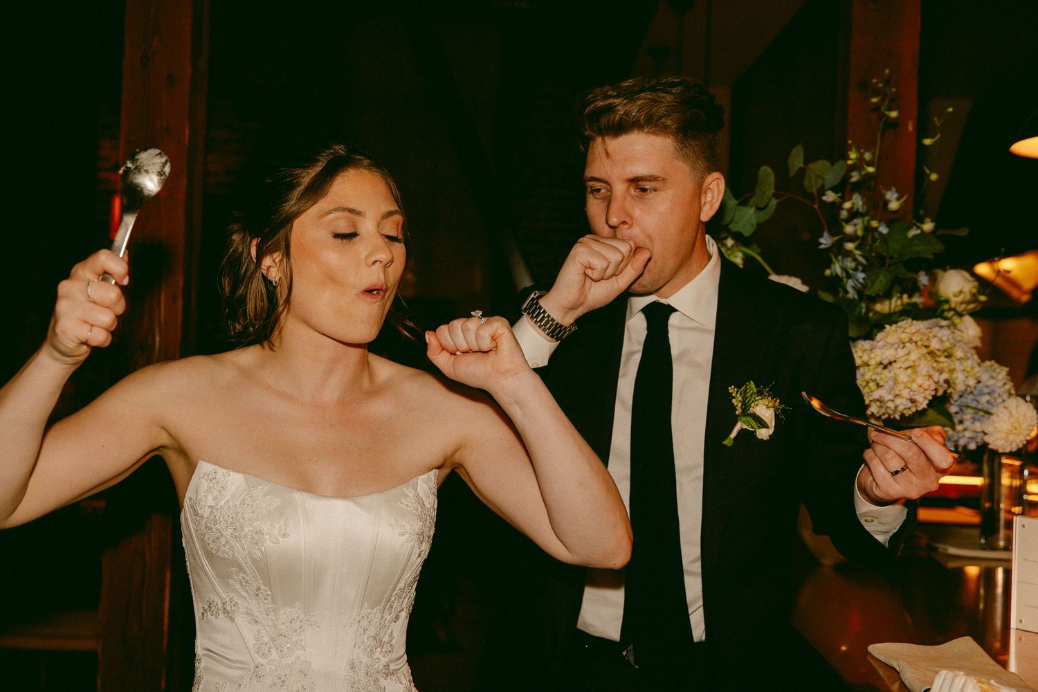 Bride and groom licking spoons and tasting wedding cake at their reception celebration.