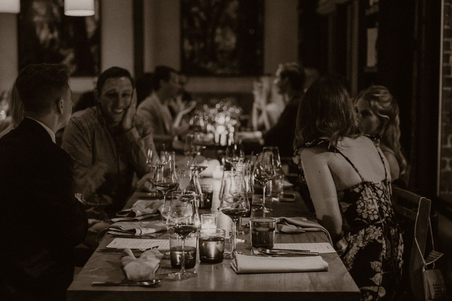 Guests enjoying an elegant candlelit dinner party at a long restaurant table in a moody, dimly lit dining room.
