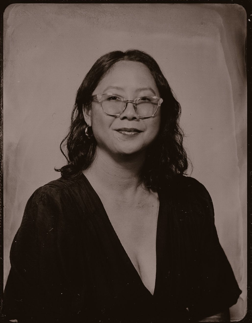 Sepia-toned tintype portrait of a smiling woman with glasses and dark wavy hair wearing a black top.