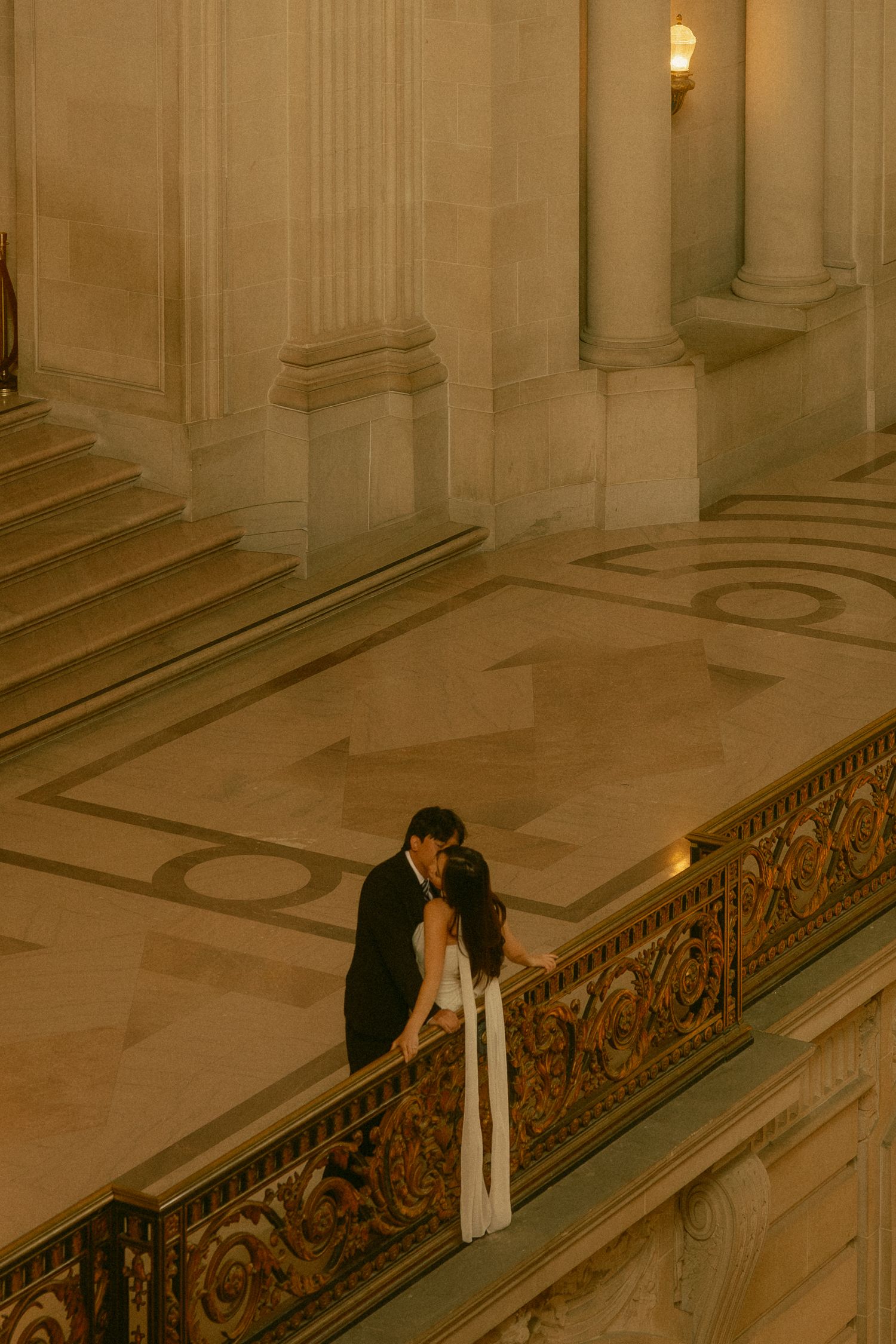 Couple sharing a romantic kiss on an ornate grand staircase railing in a golden-lit neoclassical building.
