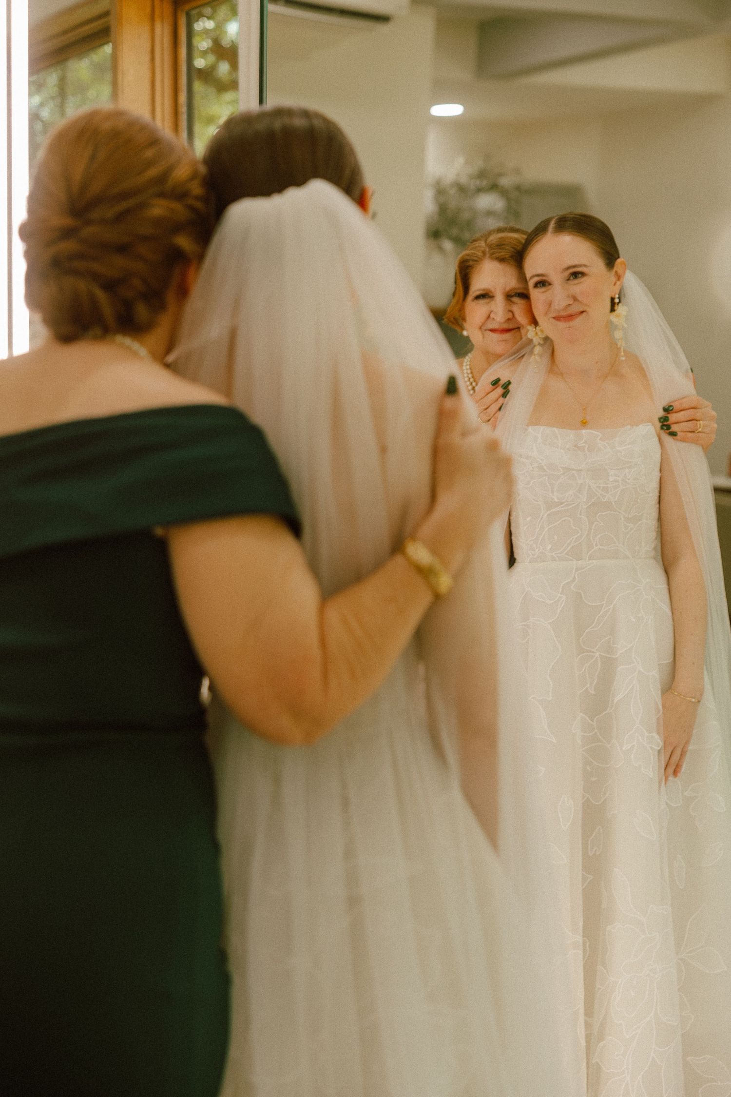 Bride in lace wedding gown and veil smiles as family members help her prepare before the ceremony.