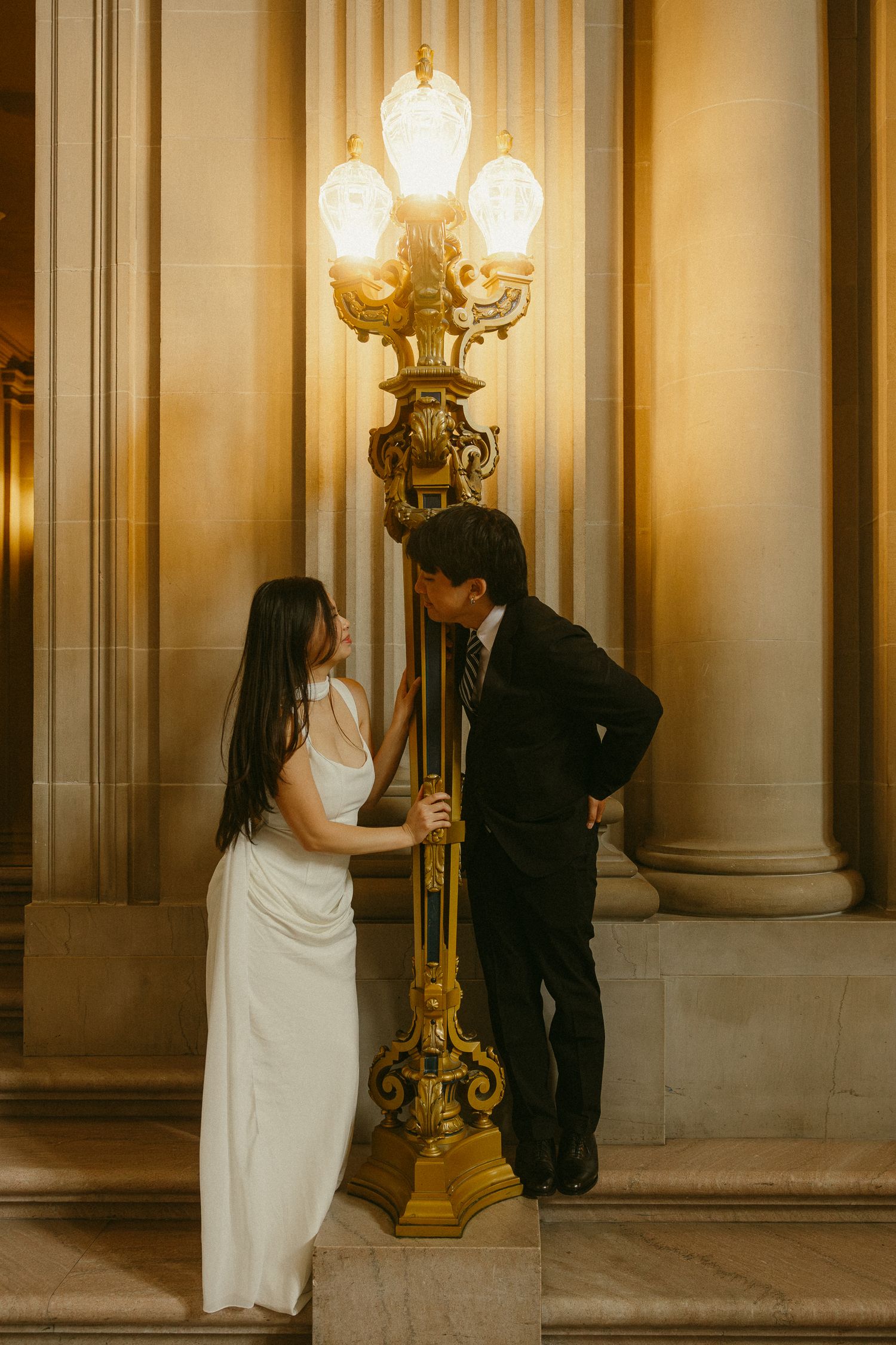 Couple in wedding attire share a romantic moment beside an ornate gold lamp post in an elegant columned hall.