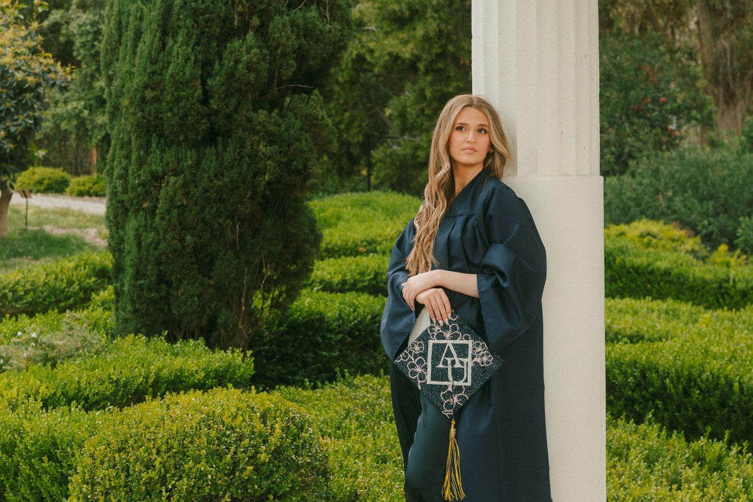 Woman in graduation gown and cap holding diploma, posing by white column in lush green garden.