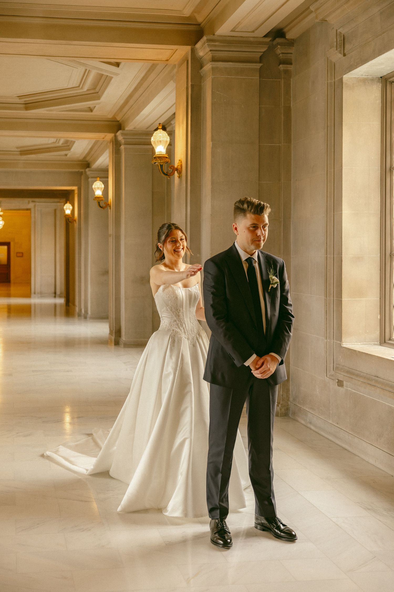 Bride in white gown taps groom's shoulder during first look in elegant marble hallway with ornate columns.