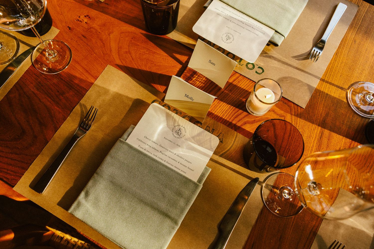 Elegant wedding reception table setting with linen napkins, amber glassware, and candlelight on warm wood surface.