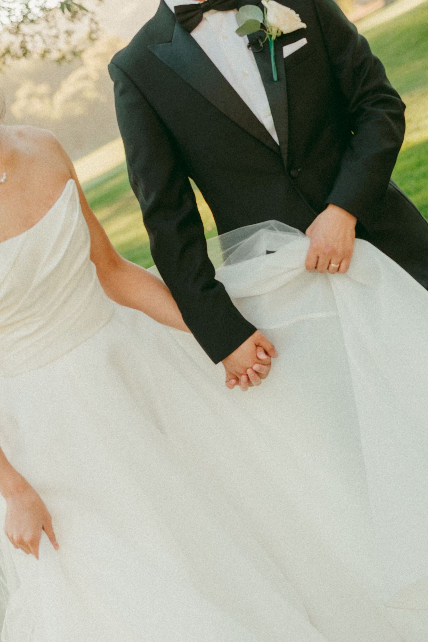 Bride and groom holding hands, wearing a white wedding gown and black tuxedo with boutonniere, walking outdoors.