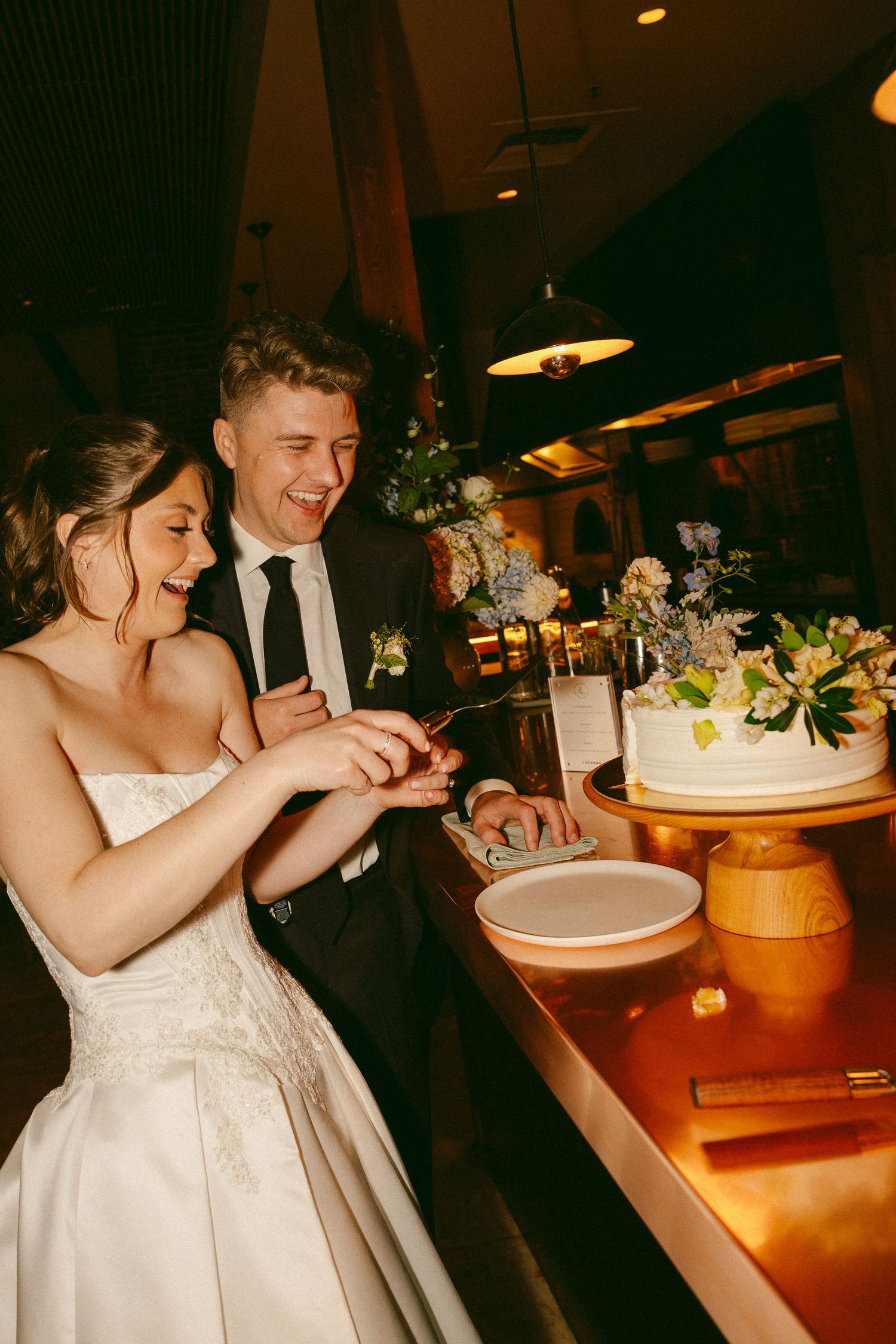 Bride and groom laughing while cutting their white wedding cake together at a warmly lit reception venue.