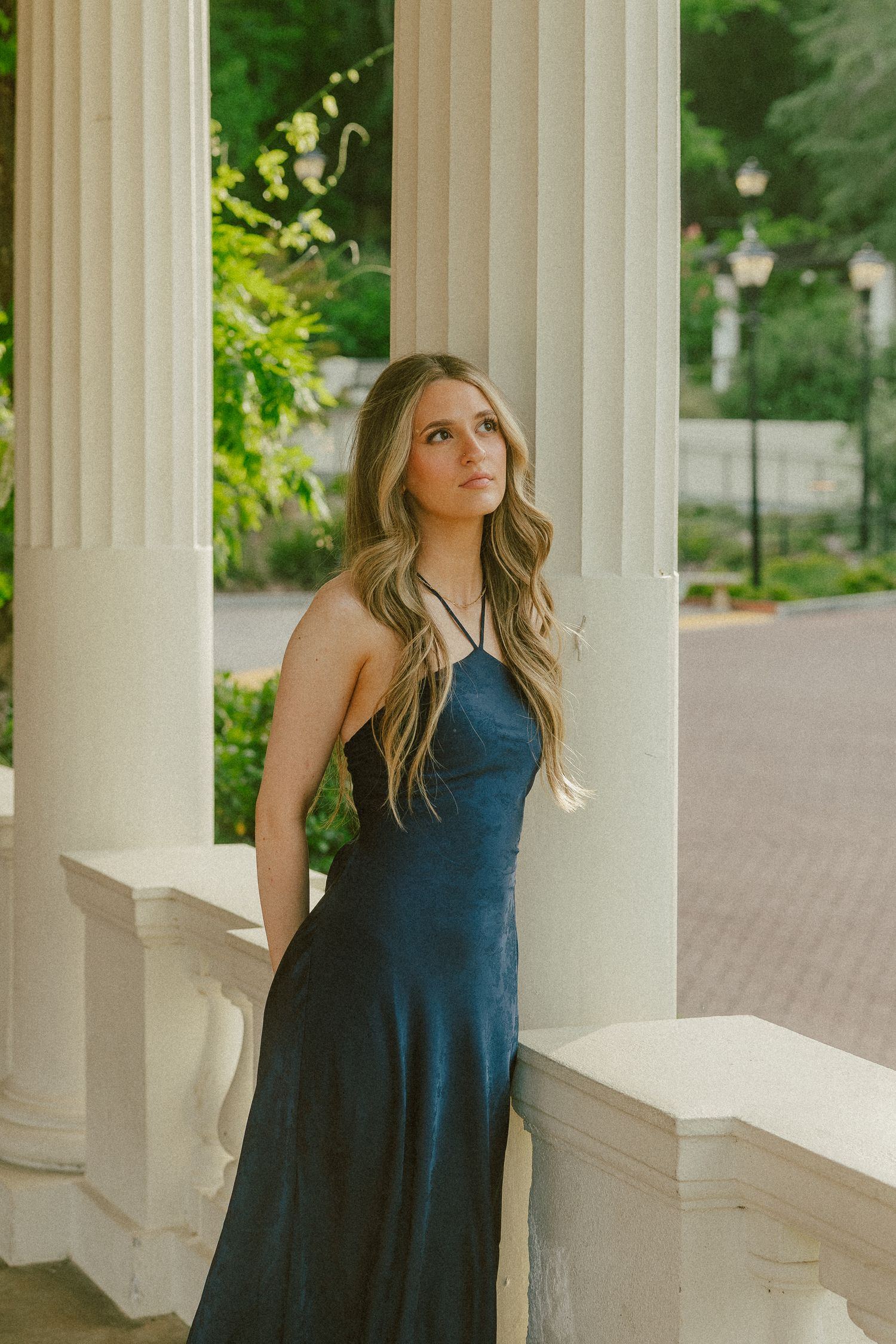 Young woman in navy blue satin dress poses gracefully between white columns in an elegant outdoor setting.