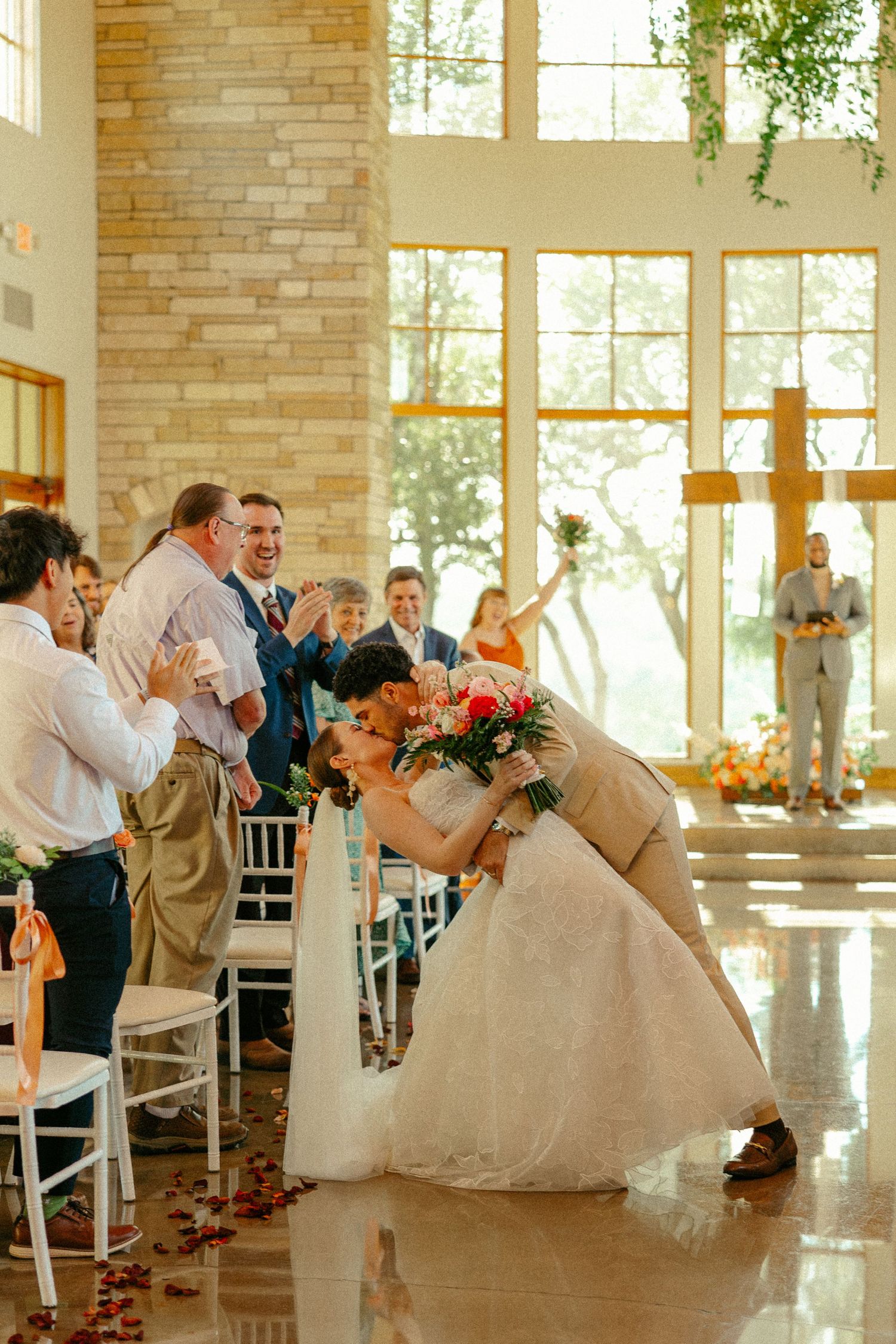 Groom dips bride for a kiss at wedding ceremony aisle in elegant chapel with large windows and cross.