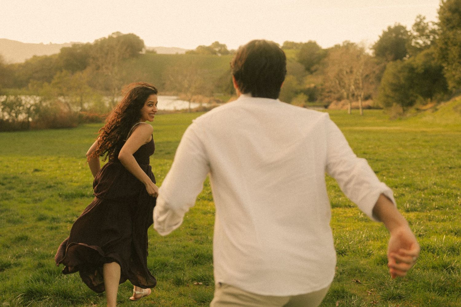 A woman in a black dress and a man in white shirt running toward each other on a green meadow at sunset.