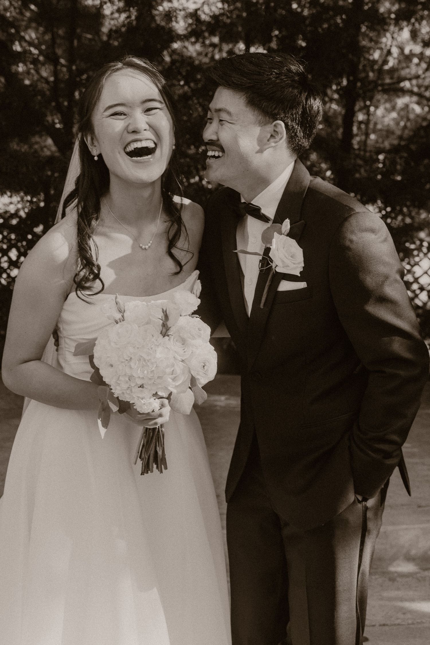Black and white photo of a laughing bride holding bouquet beside groom in tuxedo at wedding ceremony.