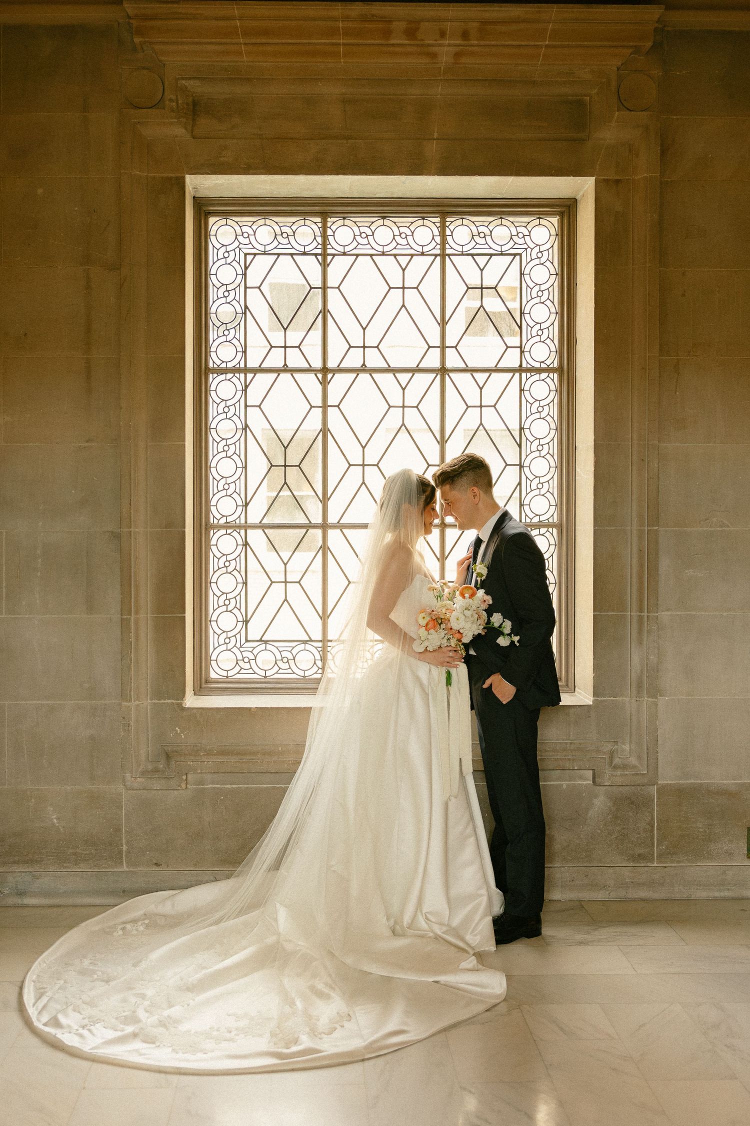 Bride and groom share a romantic moment in front of a stunning stained glass window, bride in a flowing white gown with long veil.