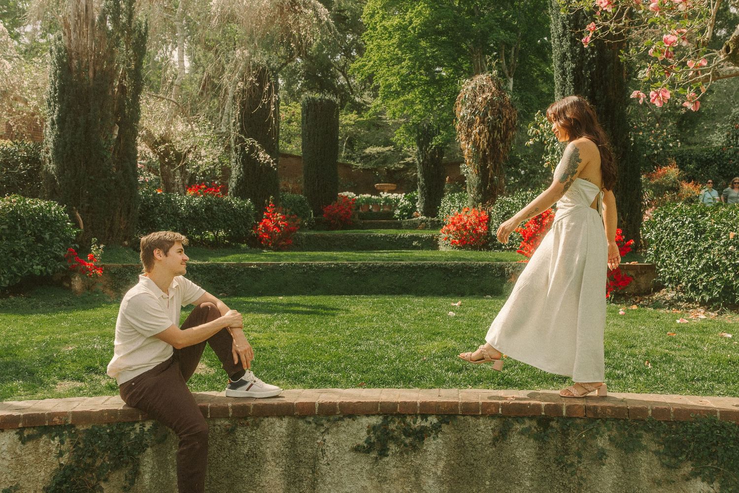 A young couple shares a romantic moment in a lush garden with colorful flowers, the woman in a white dress.